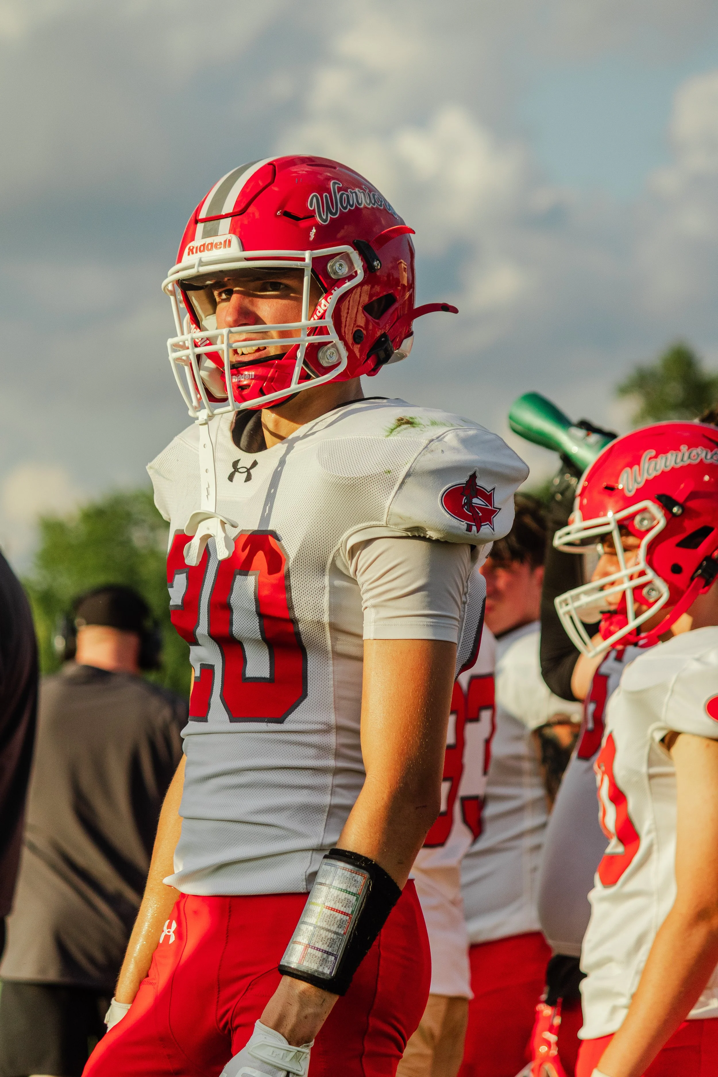 A football player wearing a red helmet with a gray and white stripe, a white jersey with red and gray numbers, and red pants, holding a play sheet in his left hand, standing on a football field with other players in similar uniforms and a cloudy sky in the background.