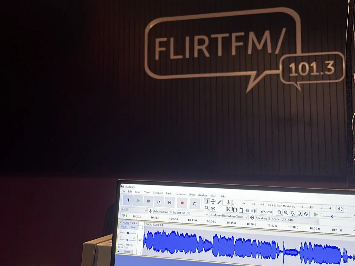 A computer screen with a waveform in an audio editing software, and a dark wall with a sign that reads 'FLIRTFM/' and '101.3' in a speech bubble.