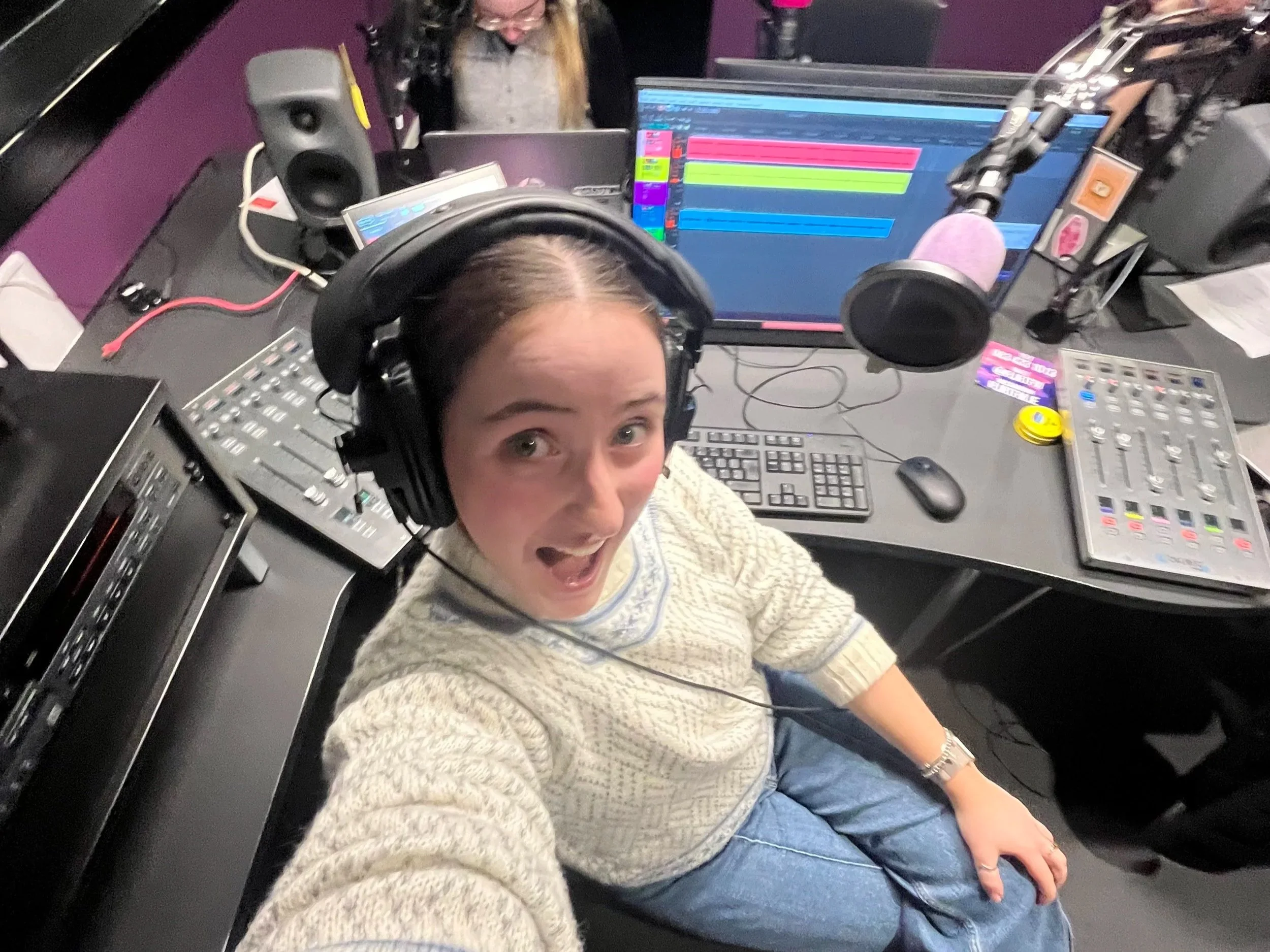 A woman in a cream-colored sweater and blue jeans is sitting in a recording studio with headphones on. She is looking up at the camera and smiling. The studio has audio equipment, a computer monitor with colorful waveforms, and a purple wall in the b
