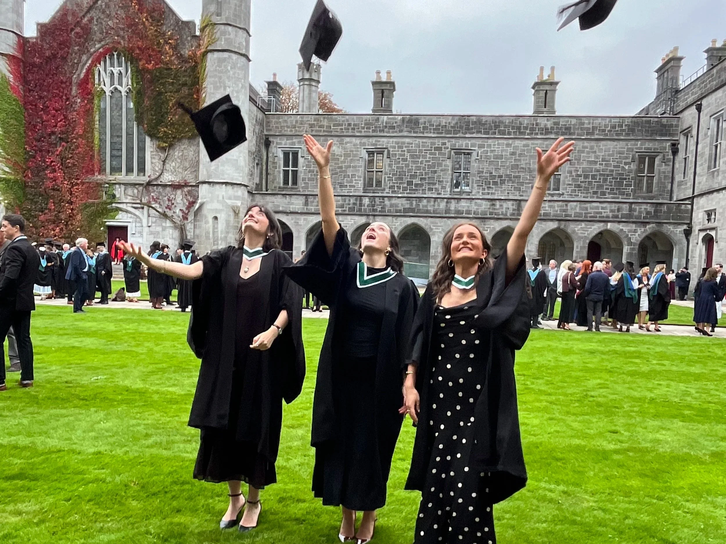 Three women in graduation gowns throwing caps into the air on a college campus with a stone building in the background.