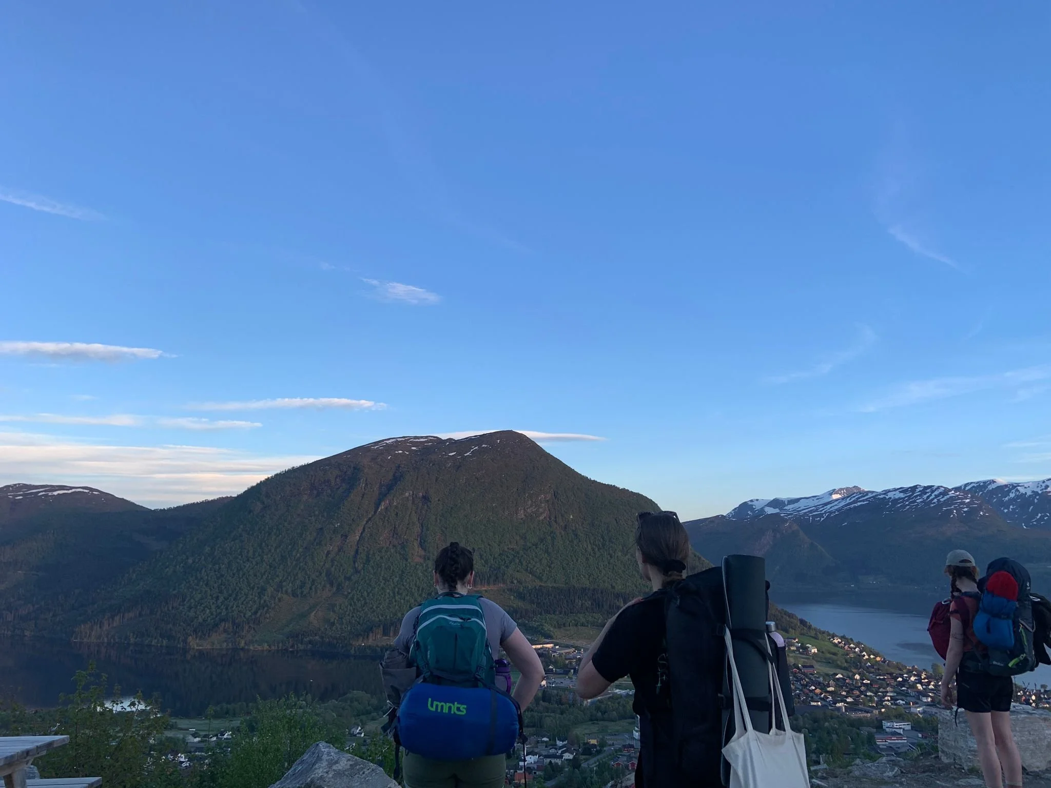 People hiking on a trail overlooking a scenic mountain landscape with green forested hills, a town, and snow-capped peaks under a clear blue sky, taken in Volda, Norway.
