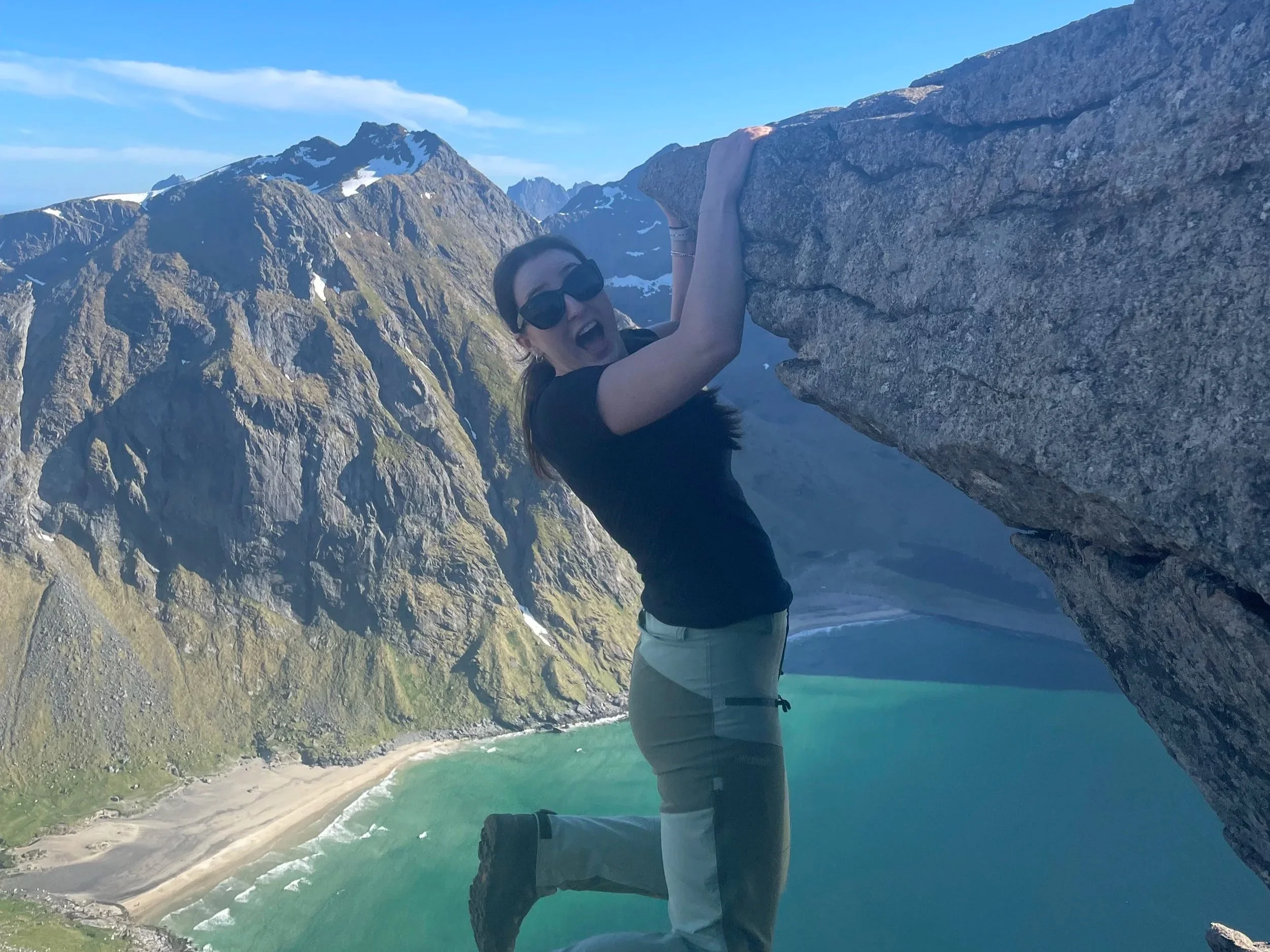 A woman with long hair wearing sunglasses, a black shirt, and gray pants climbing a large rock formation on a mountain. The background features snow-capped peaks, a green valley, and a sandy beach with turquoise water.