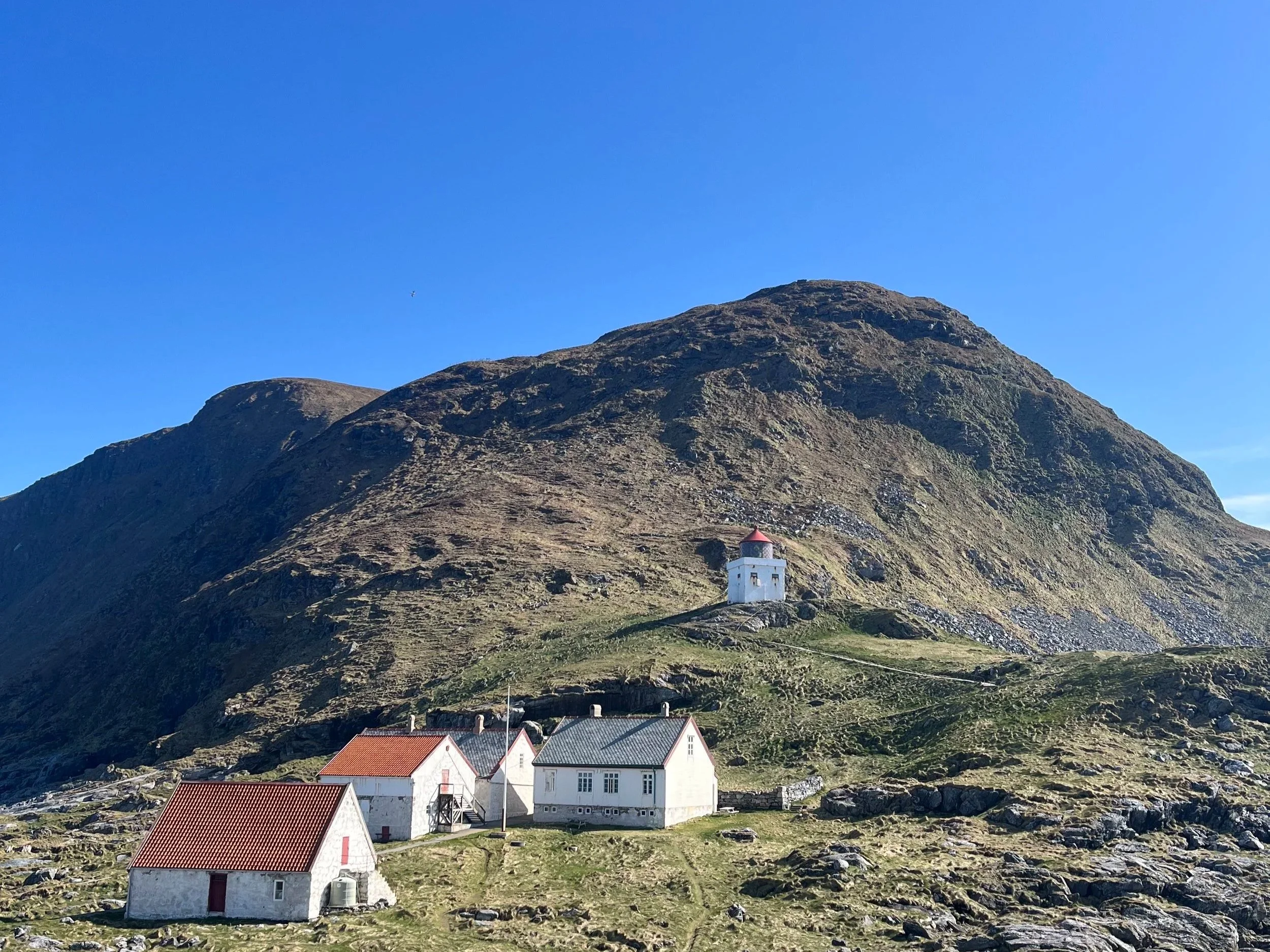 A small mountain landscape with a cluster of white houses with red roofs at the base, a white lighthouse with a red roof on the hillside, rocky terrain, and a clear blue sky, taken at Runde, Norway.
