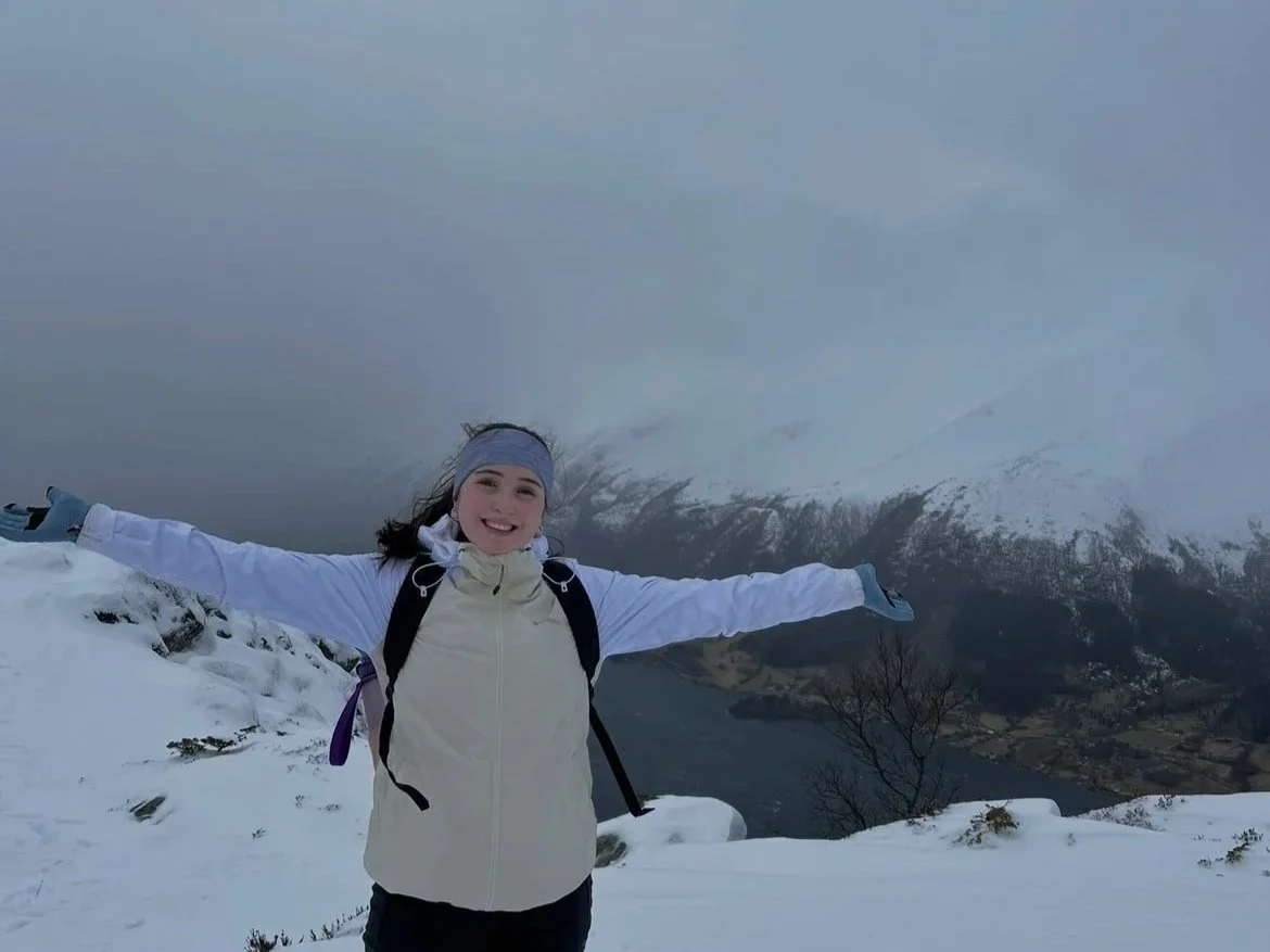 A woman standing on a snowy mountain with her arms outstretched, smiling, with clouds covering a mountain range and a lake below in the background, taken on Galten Mountain in Norway.