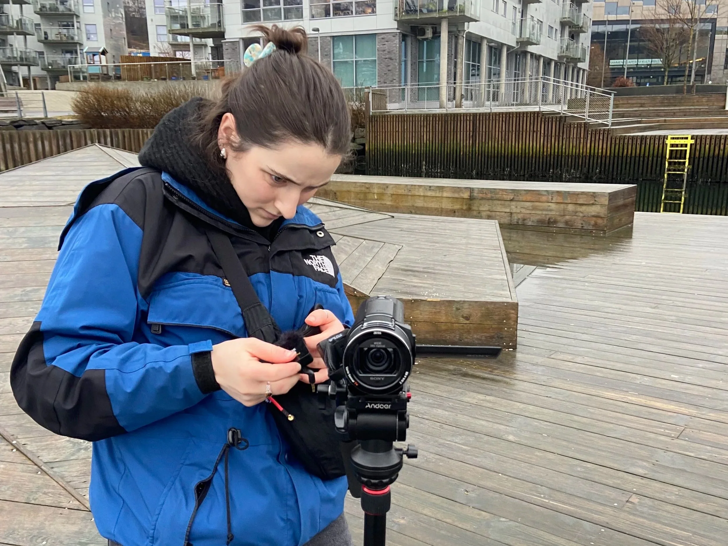 Woman in blue jacket adjusting camera mounted on tripod outdoors near modern buildings, taken in Bergen, Norway.