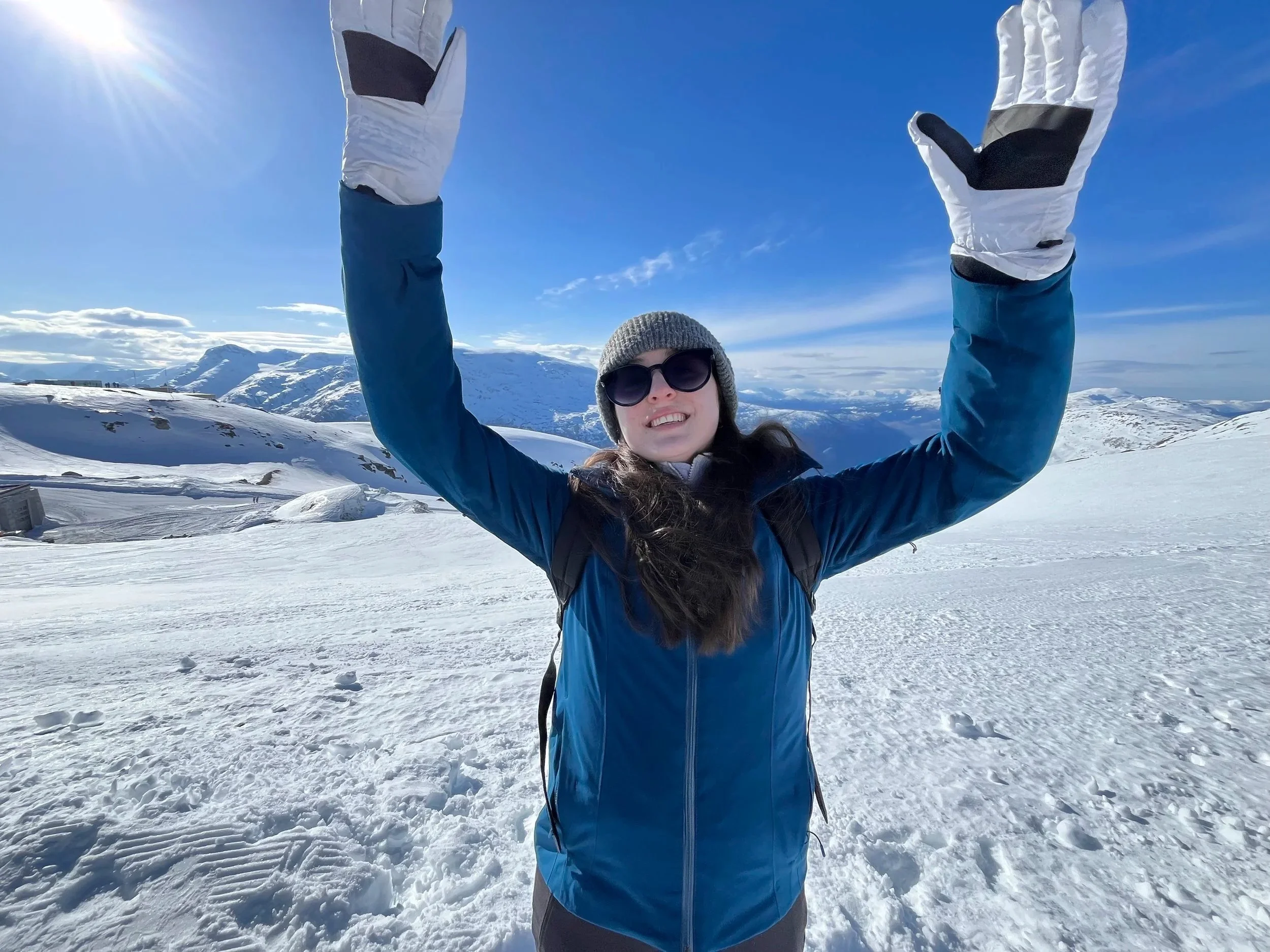 A woman wearing sunglasses, a gray beanie, a blue jacket, and white gloves is standing on a snowy mountain landscape under a clear blue sky, raising her arms in a celebratory pose.