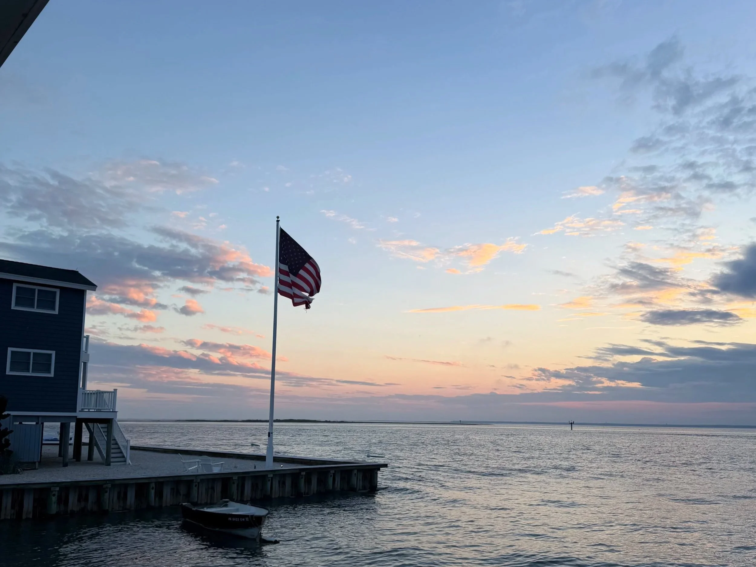 A sunset over a body of water with an American flag on a flagpole near a house and a small boat docked below. The sky has scattered clouds with warm hues, taken on Long Beach Island, NJ, America.