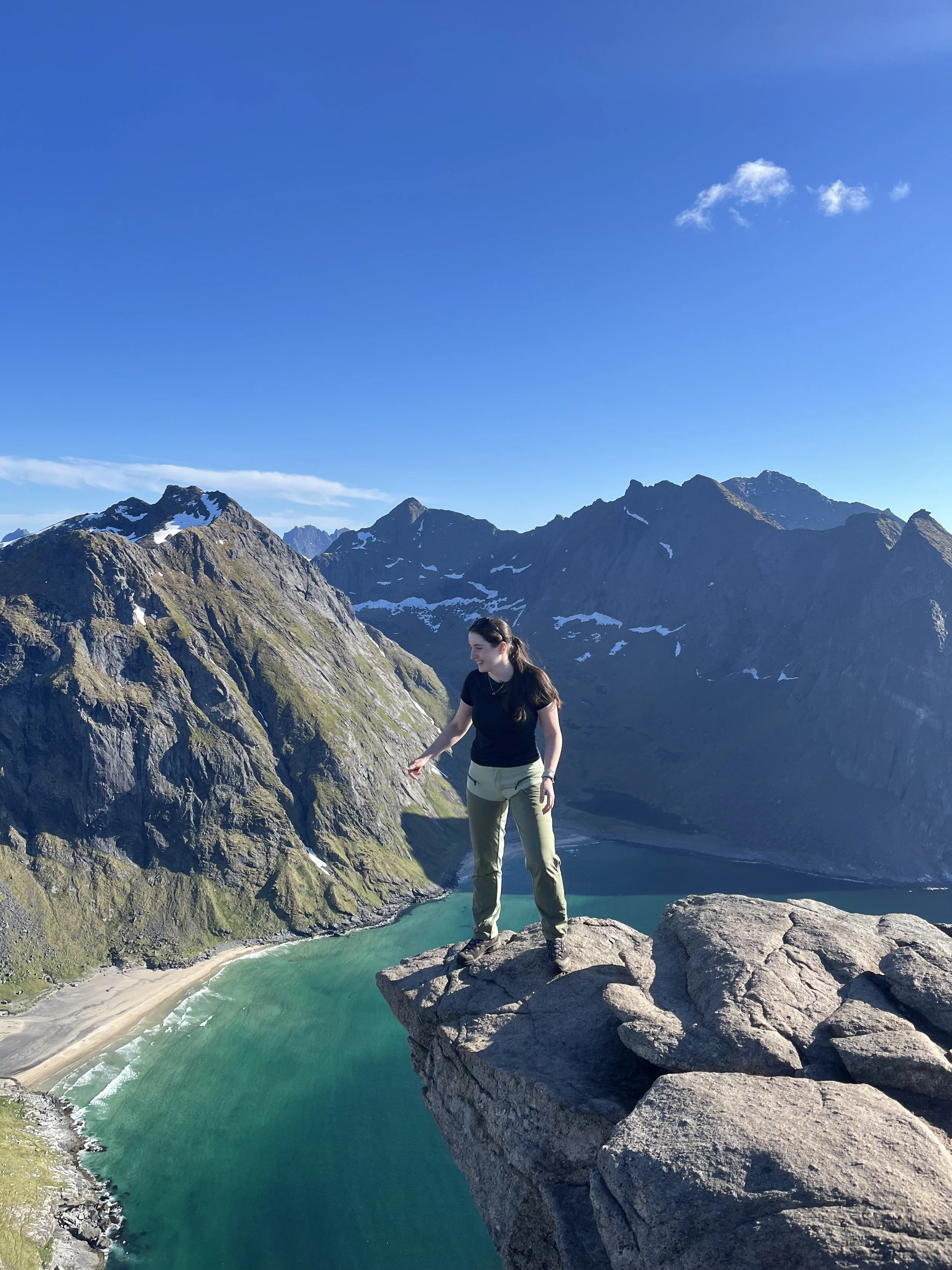 A woman standing on a rocky ledge overlooking a green river in a mountainous landscape with snow-capped peaks and a blue sky, taken at Lofotodden National Park Hike.
