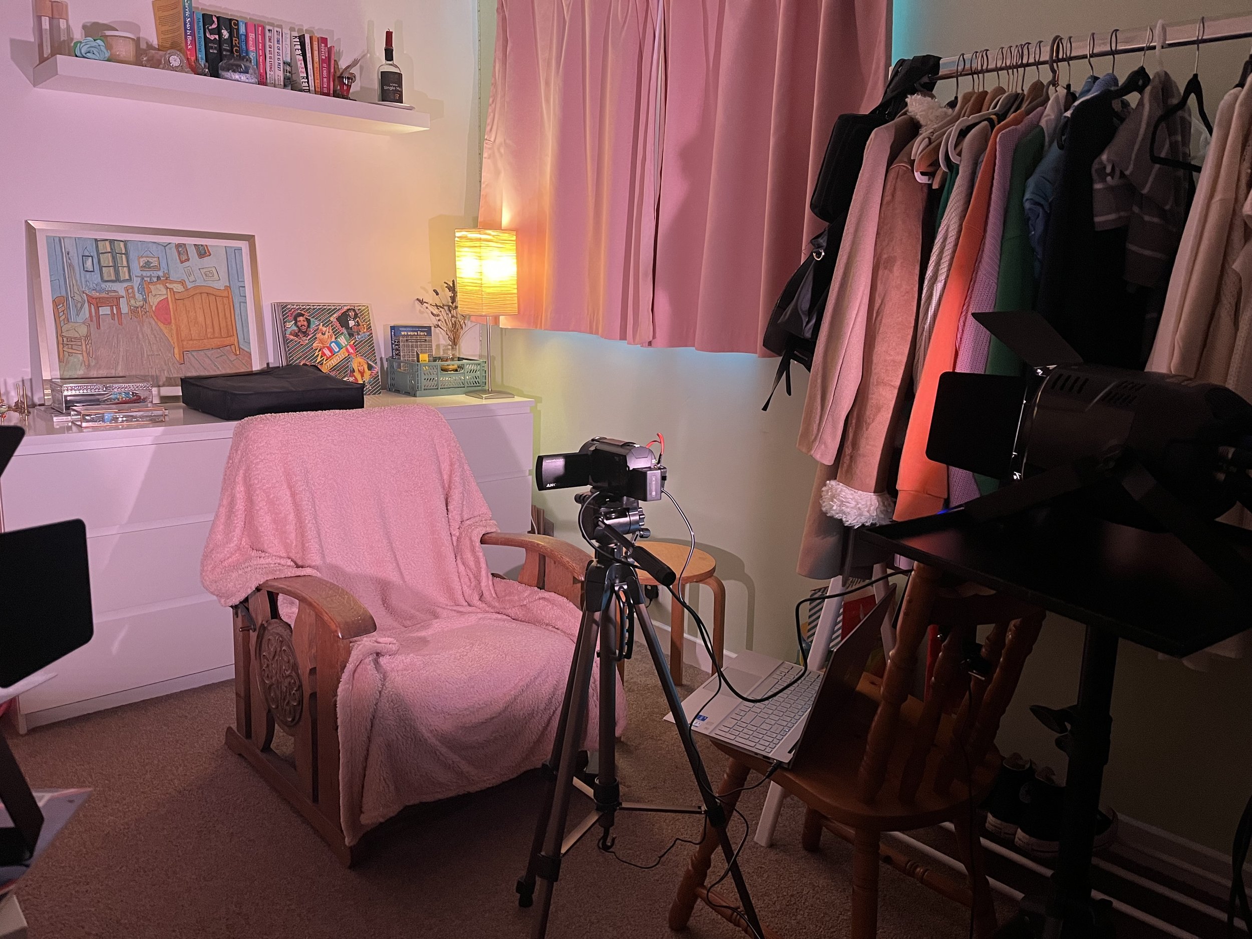 At home recording set up. There is a camera set up on a tripod in front of the chair. A cozy bedroom corner with a white dresser, framed artwork, and a pink blanket draped over a wooden chair.