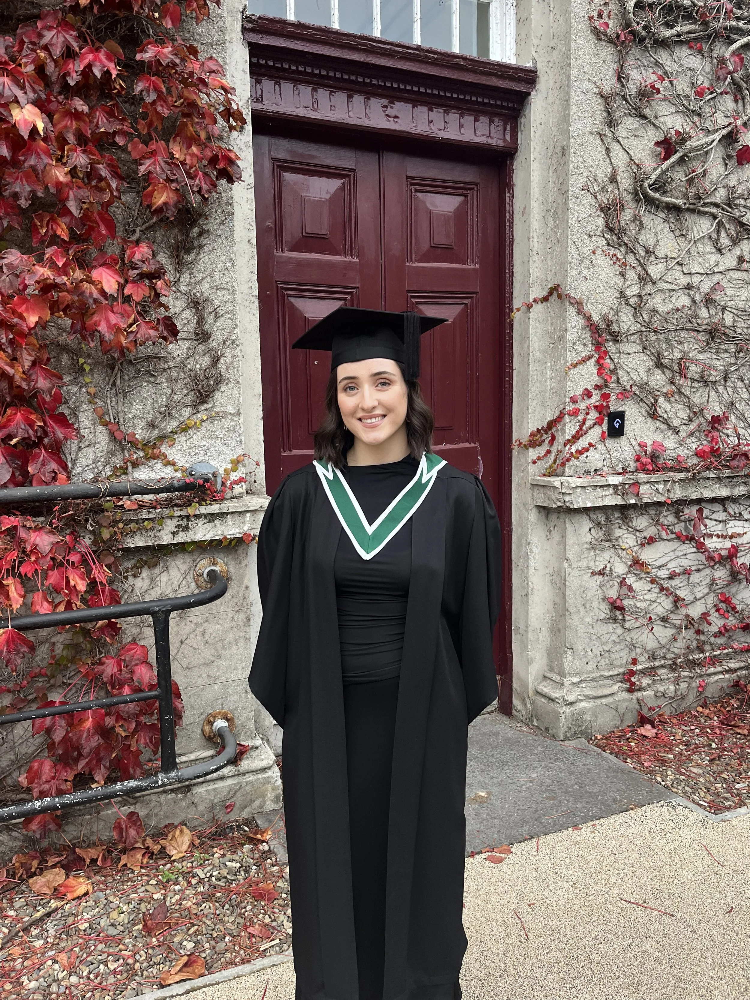 A young woman in a graduation cap and gown smiling in front of a red door with red and green autumn leaves on the wall.