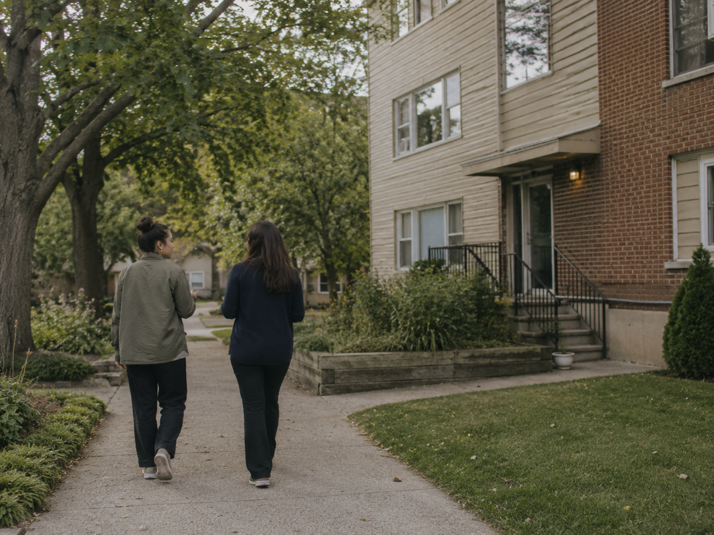Two people walking and talking outside a small apartment building