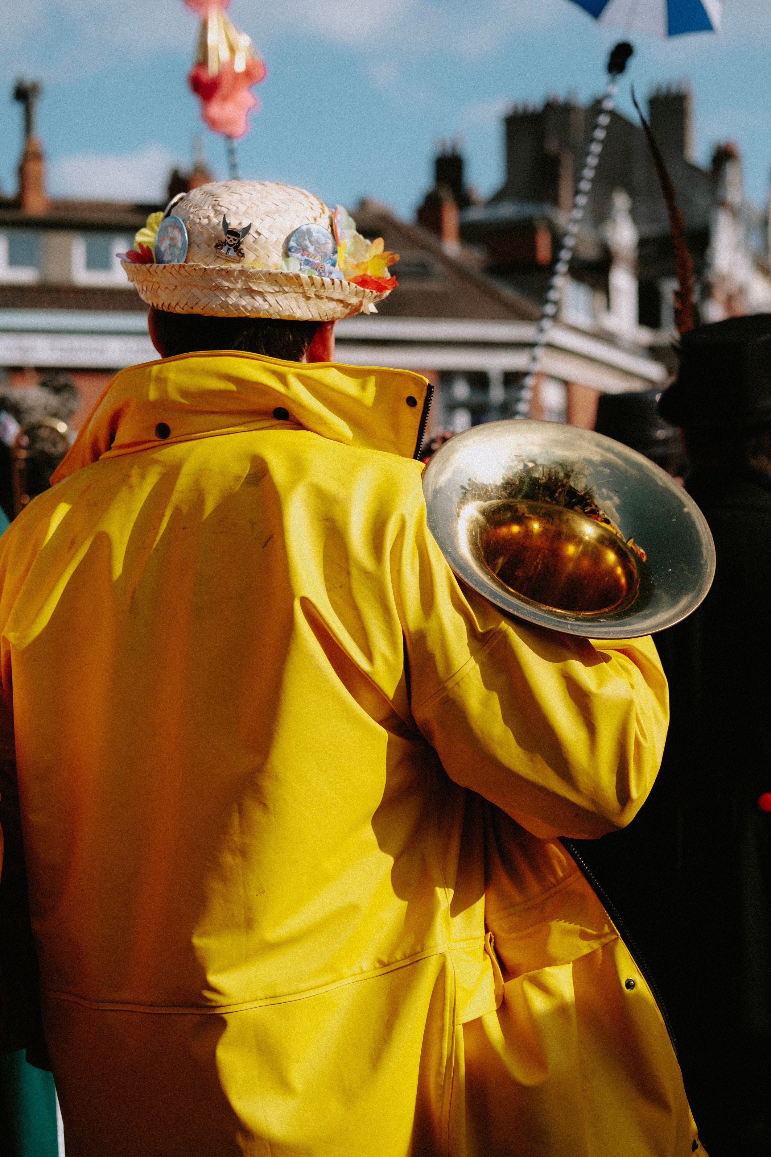 Person wearing a yellow raincoat and a straw hat decorated with playful pins, holding a shiny brass instrument in an outdoor setting with buildings and a partly cloudy sky in the background.