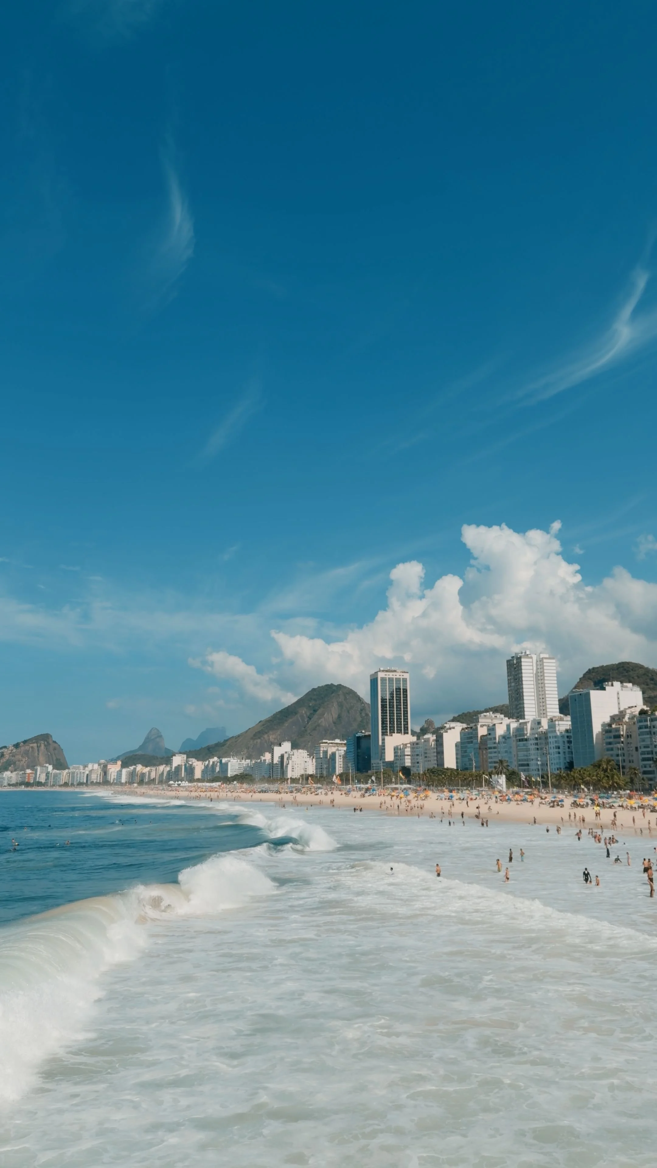 A view of a city skyline along a beach with high-rise buildings, a mountain in the background, and a blue sky with clouds.