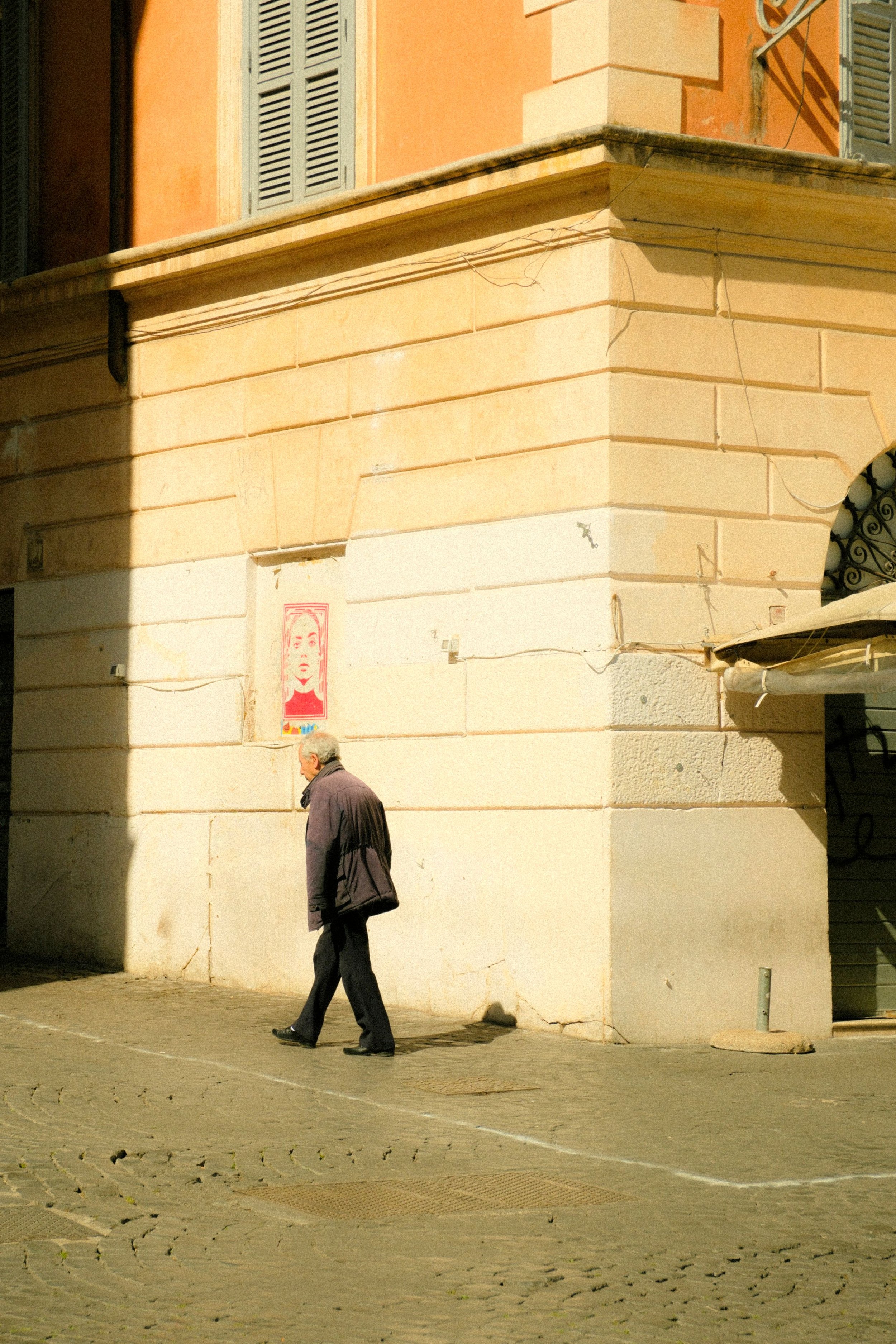 A man walking on a cobblestone street near a beige and peach building with shutters, casting a shadow on the wall.