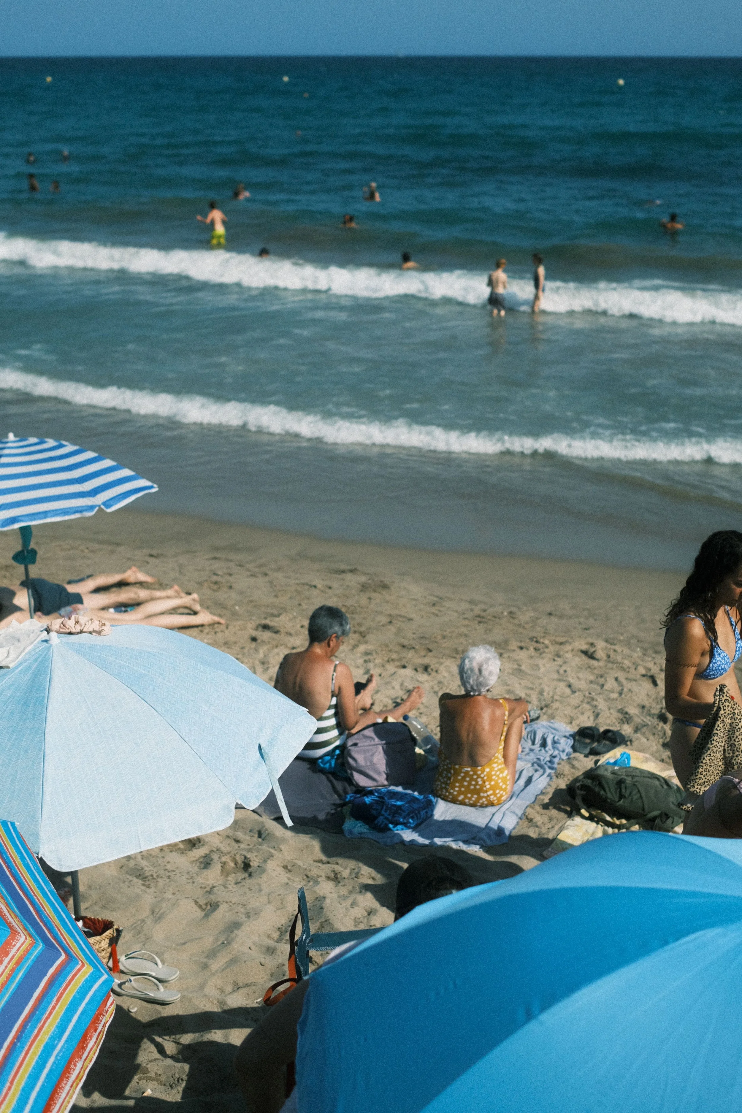People relaxing on the beach with umbrellas, and children playing in the water, with the ocean and clear sky in the background.