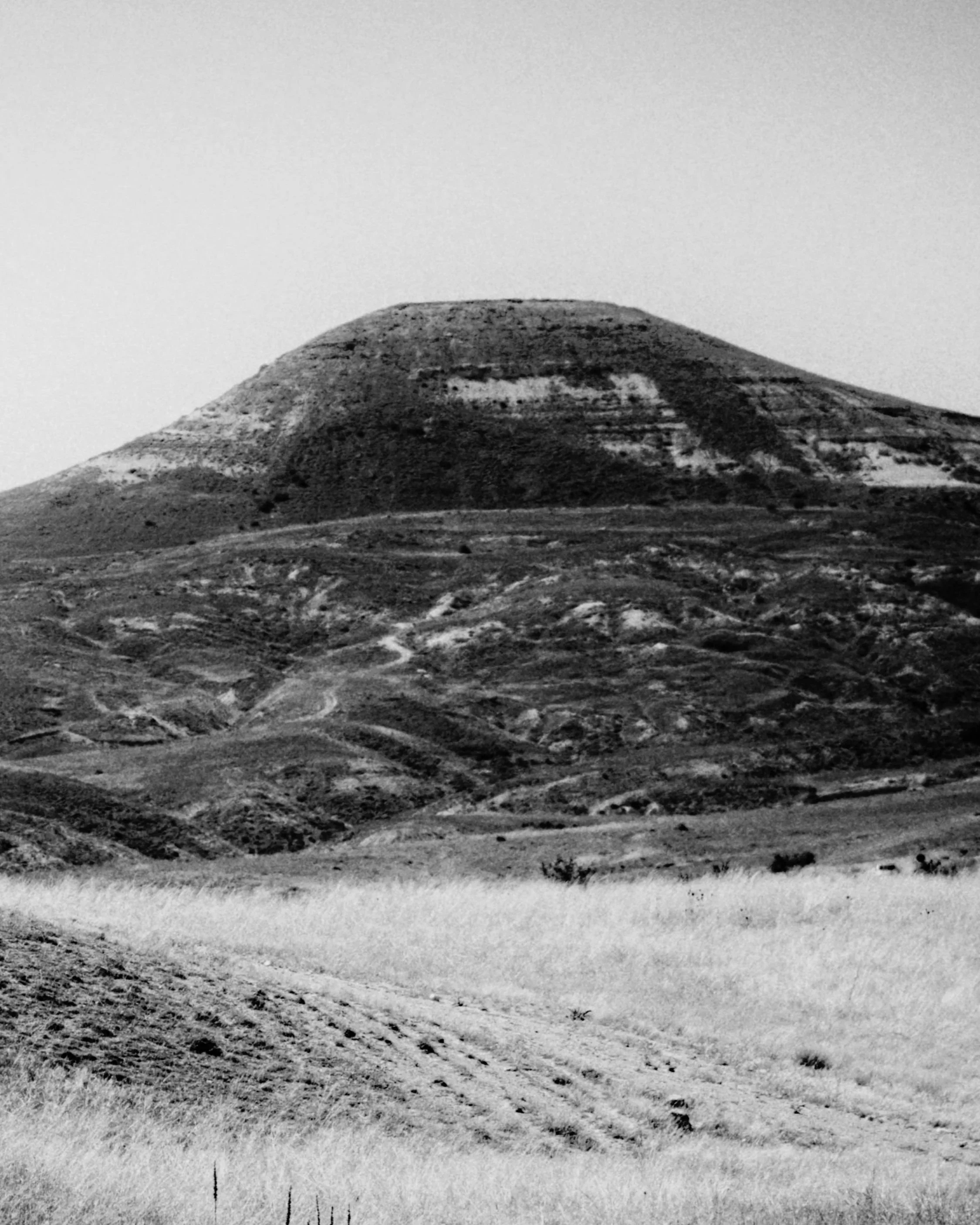 A black and white photograph of a hill or mountain with a flat top, with terrain and dirt paths in the foreground.