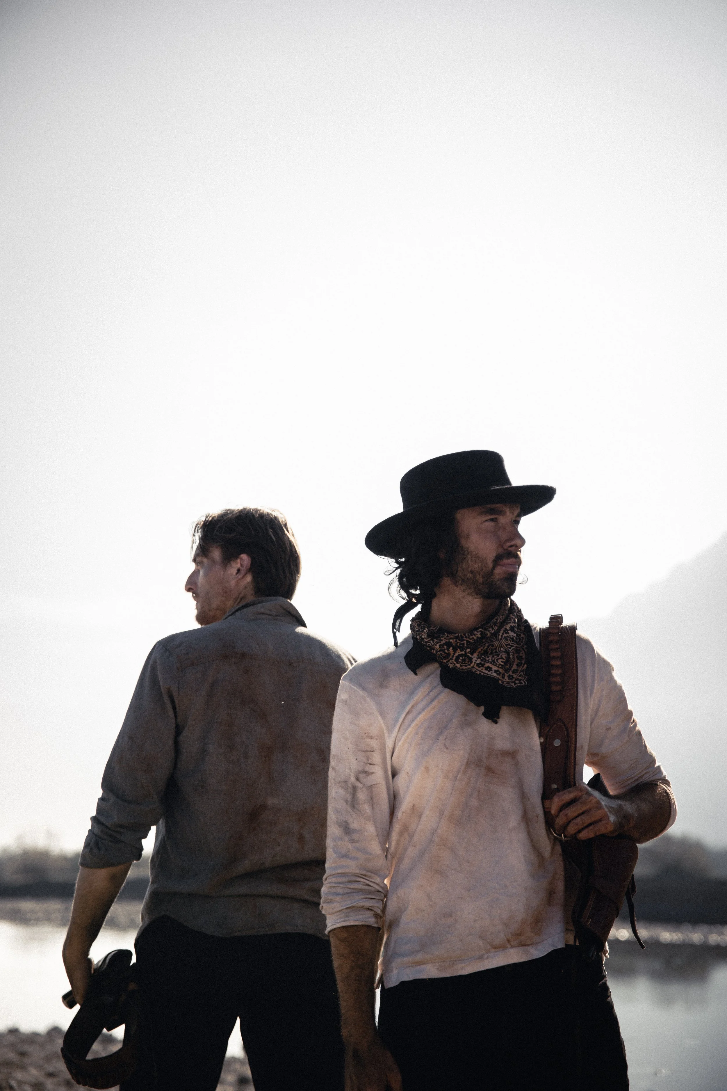 Two men standing outdoors near a body of water, one with a guitar, dressed in casual clothes including a bandana and a wide-brimmed hat, and the other with messy hair and a jacket, with the background showing a clear sky and natural landscape.