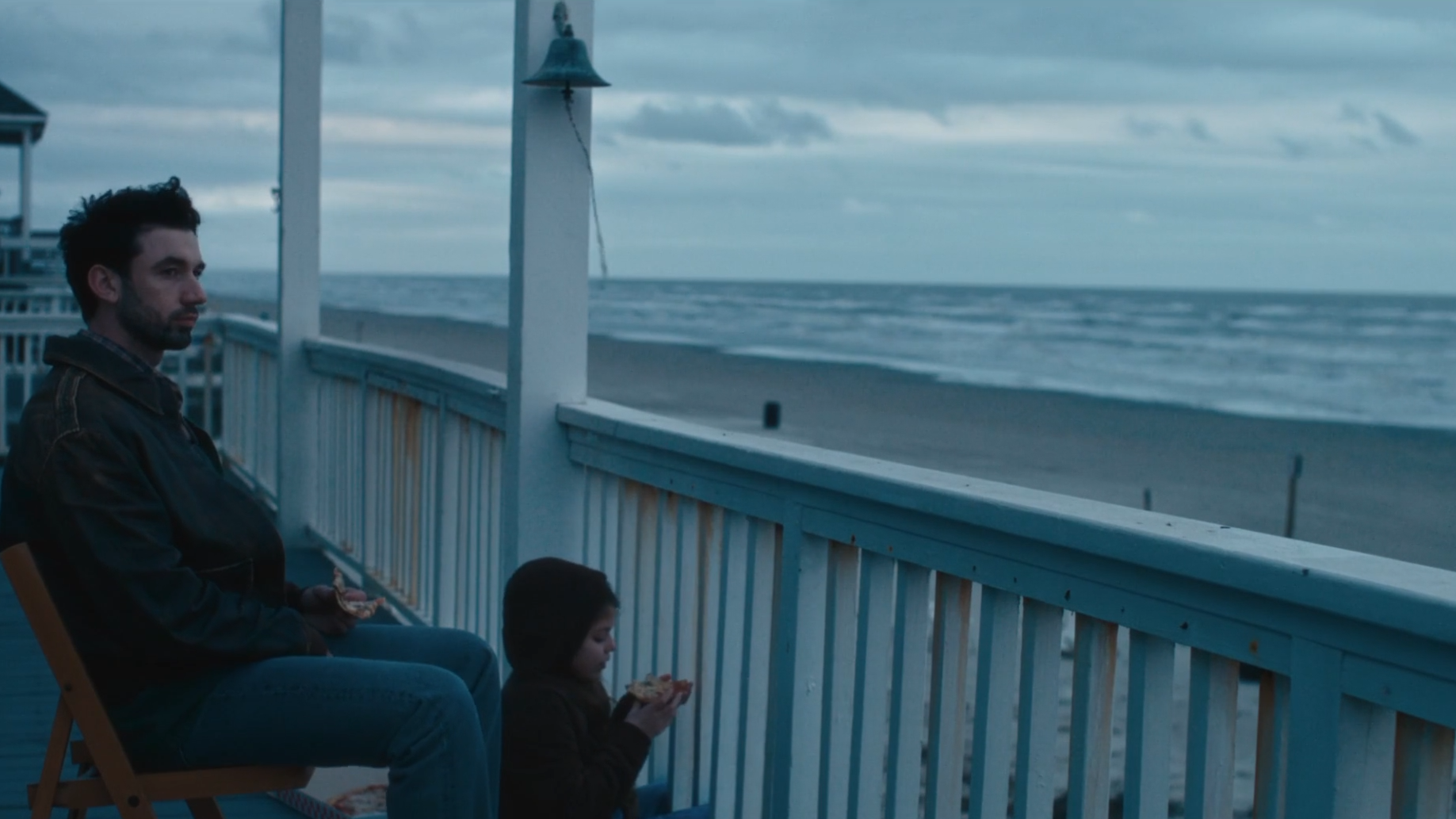 A man and two children sitting on a pier overlooking the ocean during cloudy weather, with a lighthouse bell hanging on a white railing nearby.