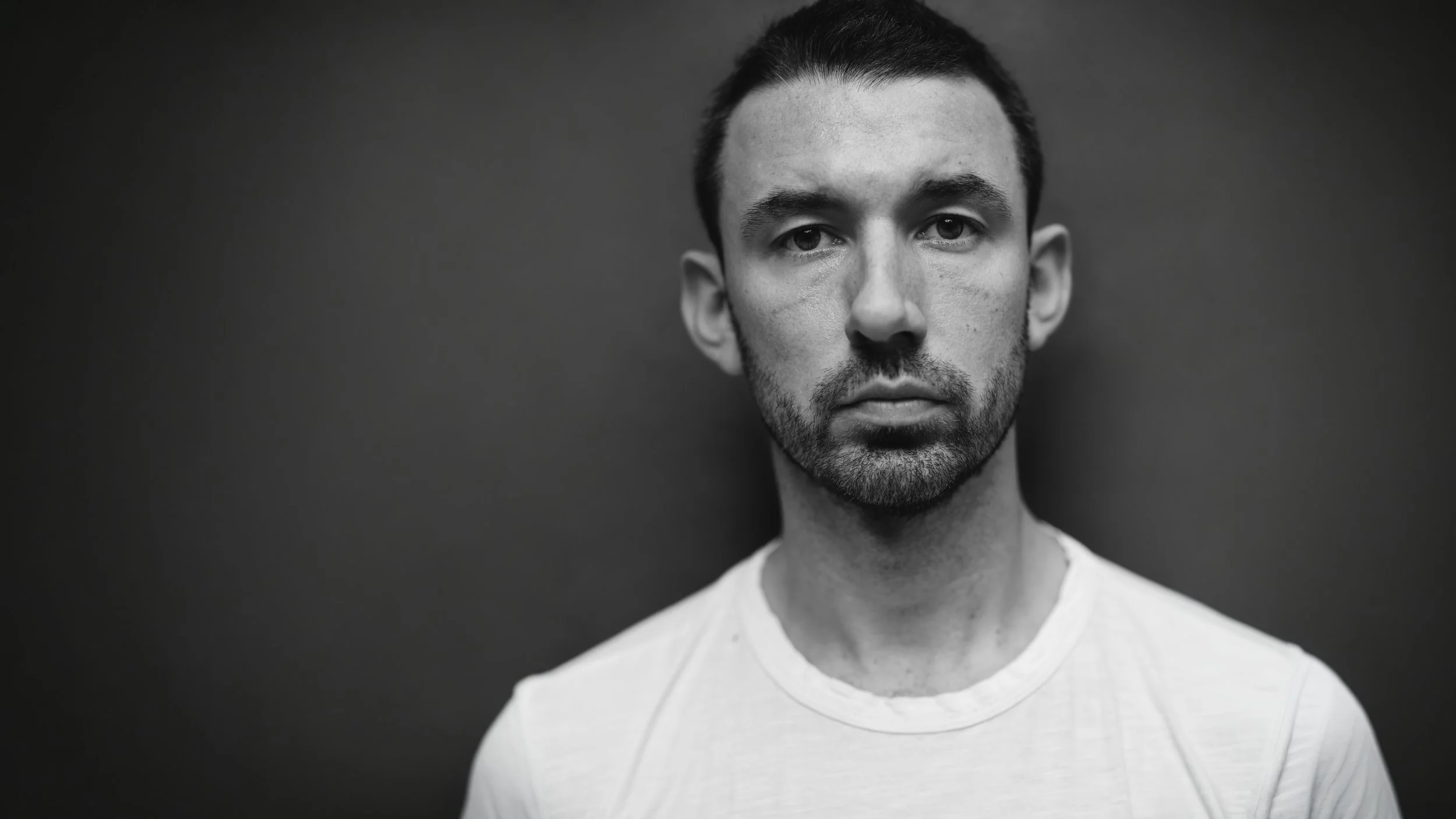 Black and white portrait of a young man with short dark hair, trimmed beard, wearing a light-colored t-shirt, against a dark background.
