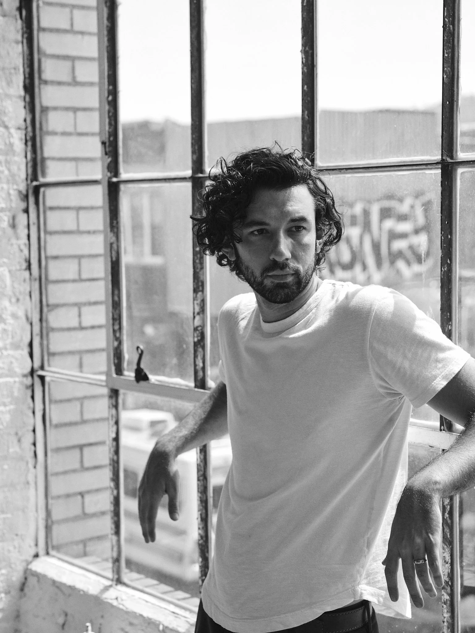 A young man with curly hair and a beard standing by a window with metal bars, looking outside on a sunny day.