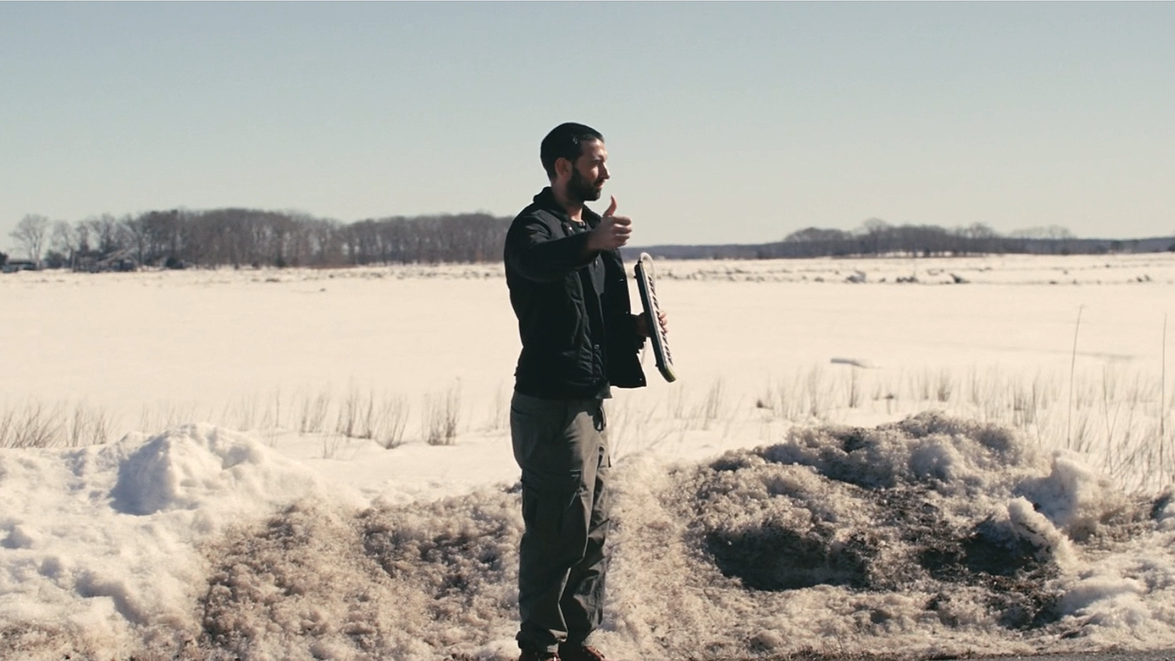 A man standing in a snowy field, holding a skateboard, giving a thumbs-up gesture.