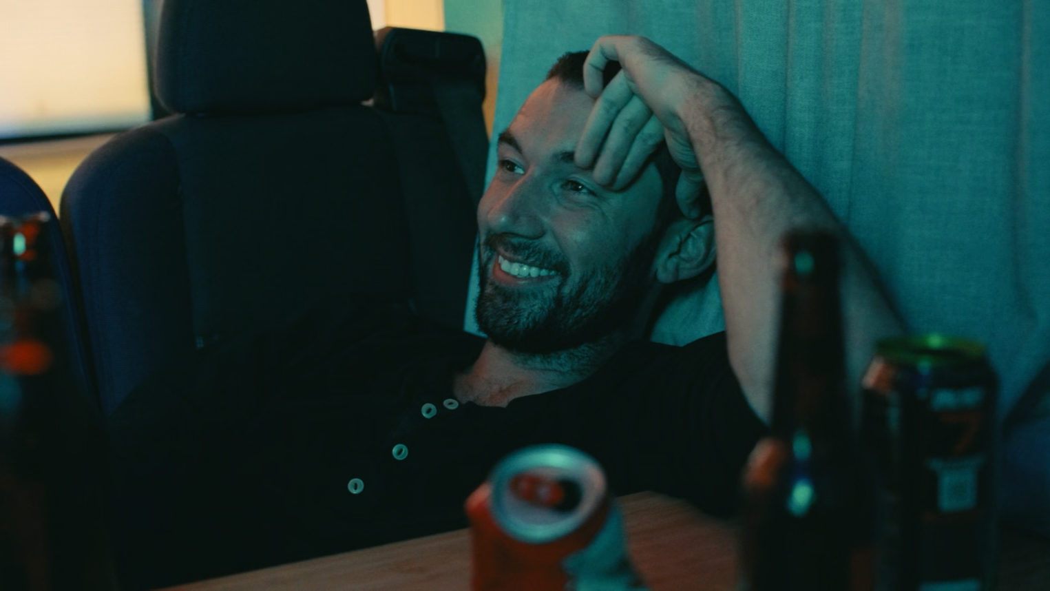 A smiling man with a beard and short hair, lying on a couch or bed with a teal wall behind him, surrounded by beer bottles and cans, in a relaxed setting.