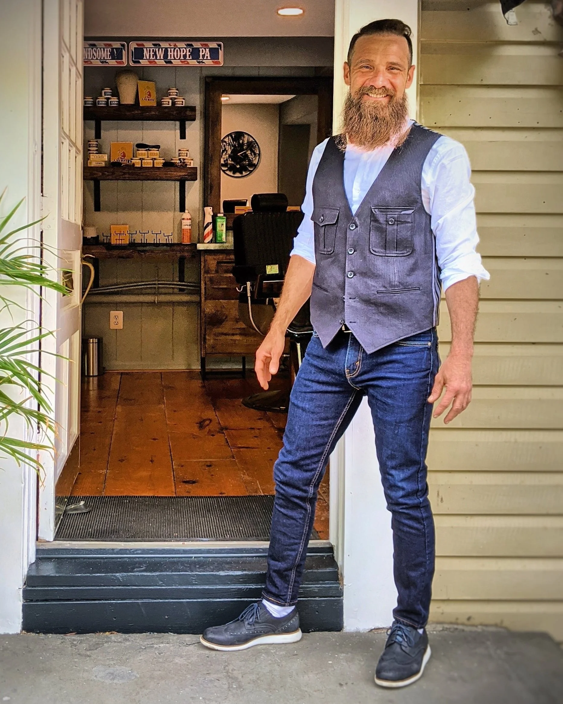 A man with a beard wearing a gray vest, white shirt with rolled-up sleeves, and dark jeans standing at the doorway of a house. The house has beige siding and a wooden step. Visible in the background are shelves with products, a black office chair, and a wall clock.