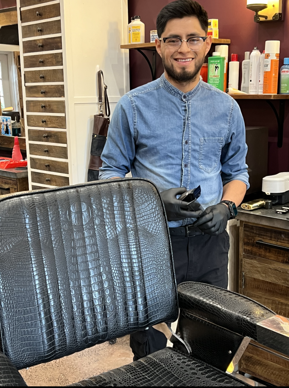 A young man with glasses and a beard, wearing a blue denim shirt and black gloves, holding a small device in a barbershop or salon. The background features shelves with hair care products and tools.