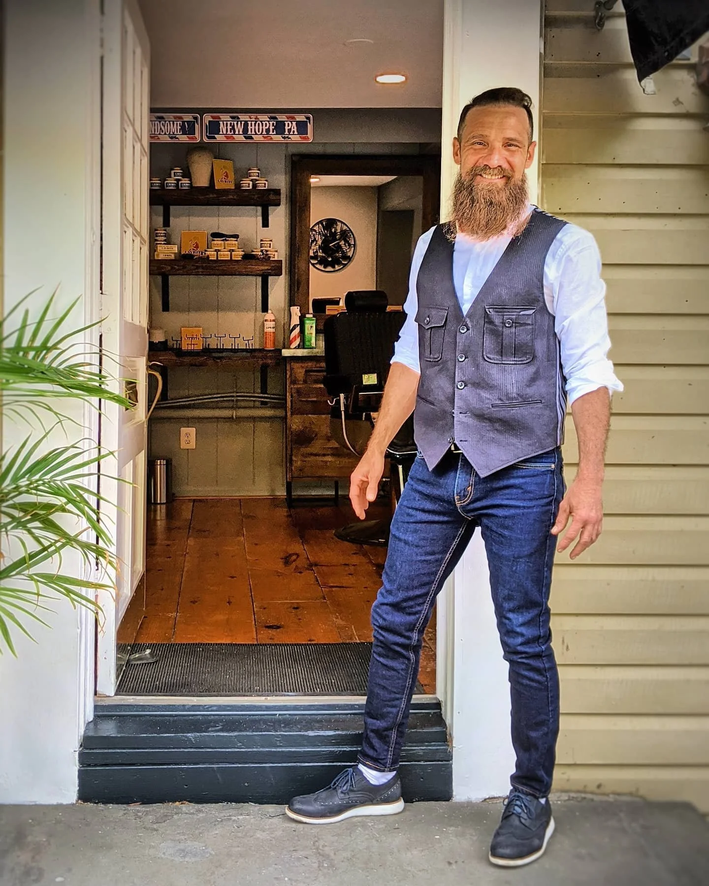 A man with a beard and mustache smiling and standing in front of a house entrance. He is wearing a white shirt, gray vest, dark jeans, and gray sneakers. The house has yellow siding and an open door leading inside, where shelves with products and signs labeled "HANDSOME" and "NEW HOPE PA" are visible.