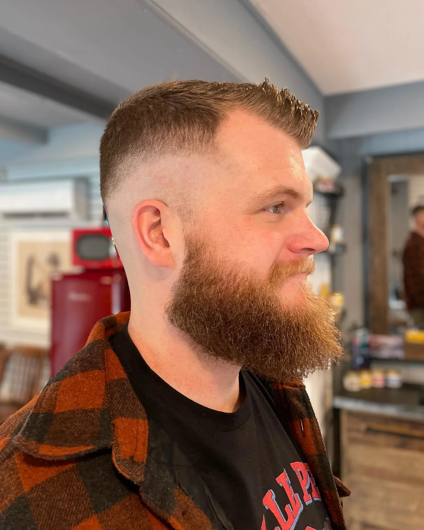 A side profile of a man with a short, neatly styled haircut and a full beard, wearing a black T-shirt and an orange and black checkered flannel shirt, in a barbershop or salon.