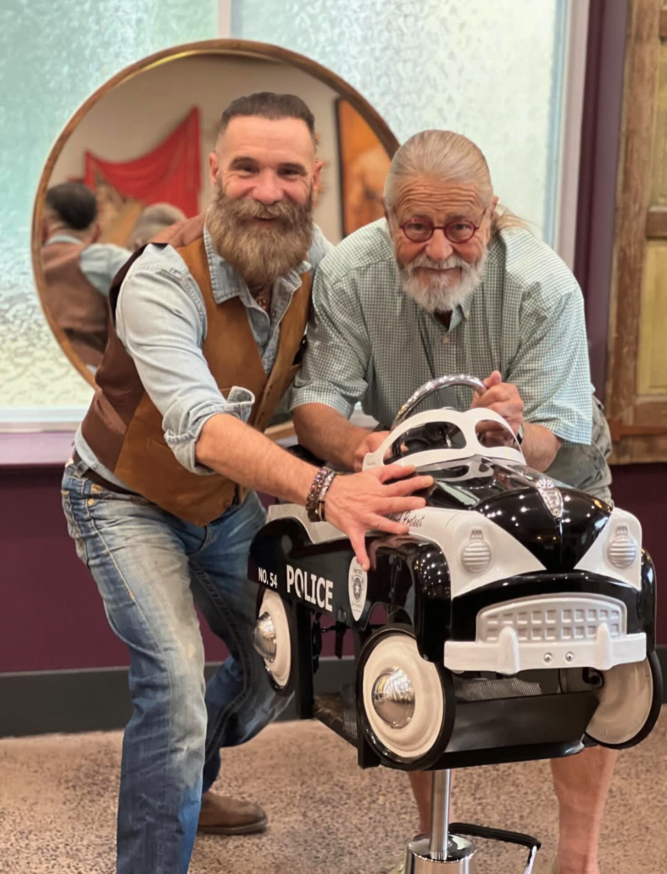 Two men, one middle-aged with a beard and another elderly with glasses, are smiling and holding a toy police car. The middle-aged man is wearing a denim shirt with a brown vest, while the elderly man is wearing a checkered shirt. Behind them is a mirror reflecting the scene and part of the room's decor.
