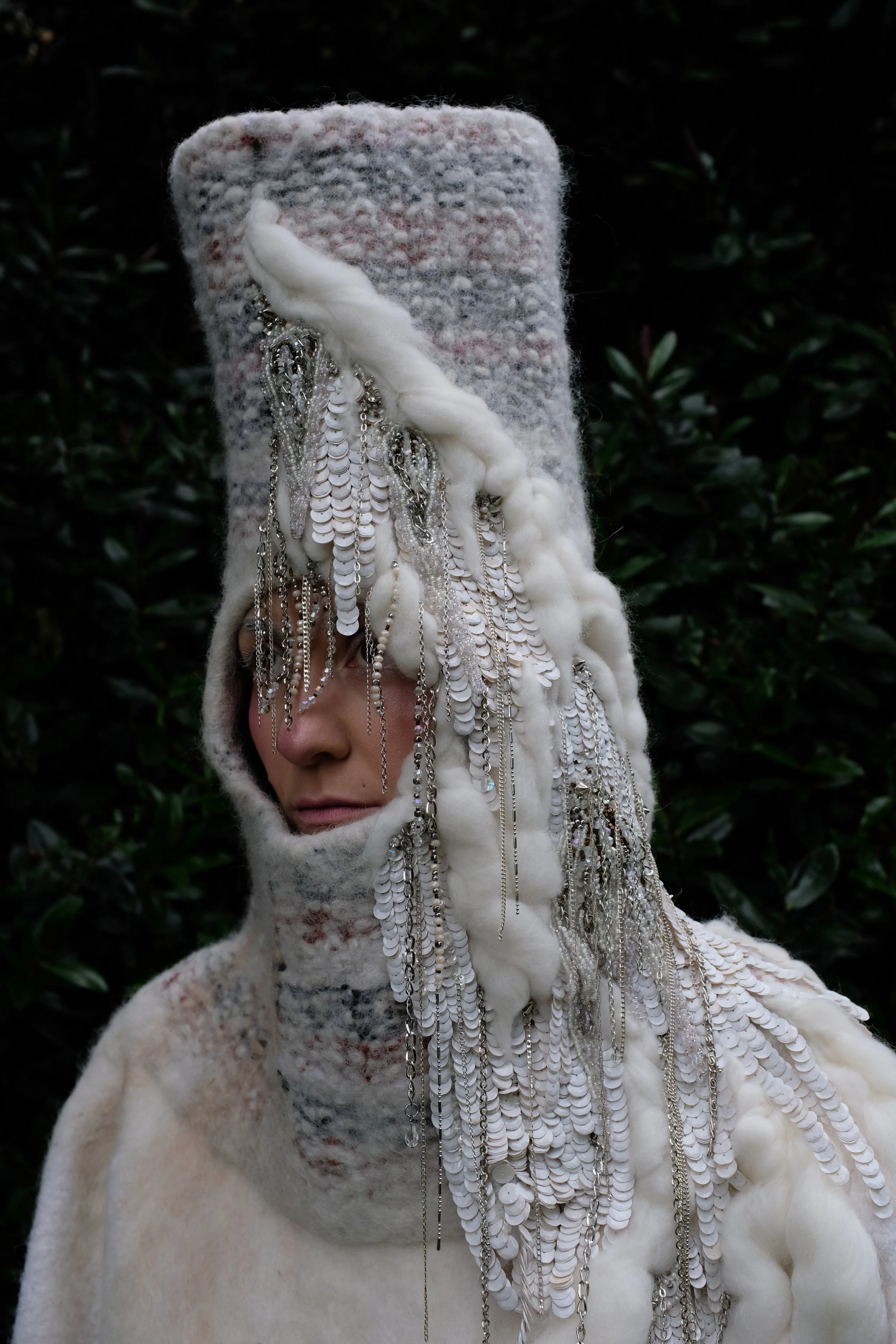 A person wearing a high, furry, multicolored hat that covers most of their face, decorated with numerous chains, beads, and sequins hanging from it and their shoulders, against a background of dark green foliage.