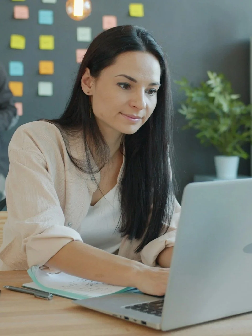 A woman with long dark hair smiling at her laptop in an office with colorful sticky notes on a dark wall behind her and a potted plant nearby.