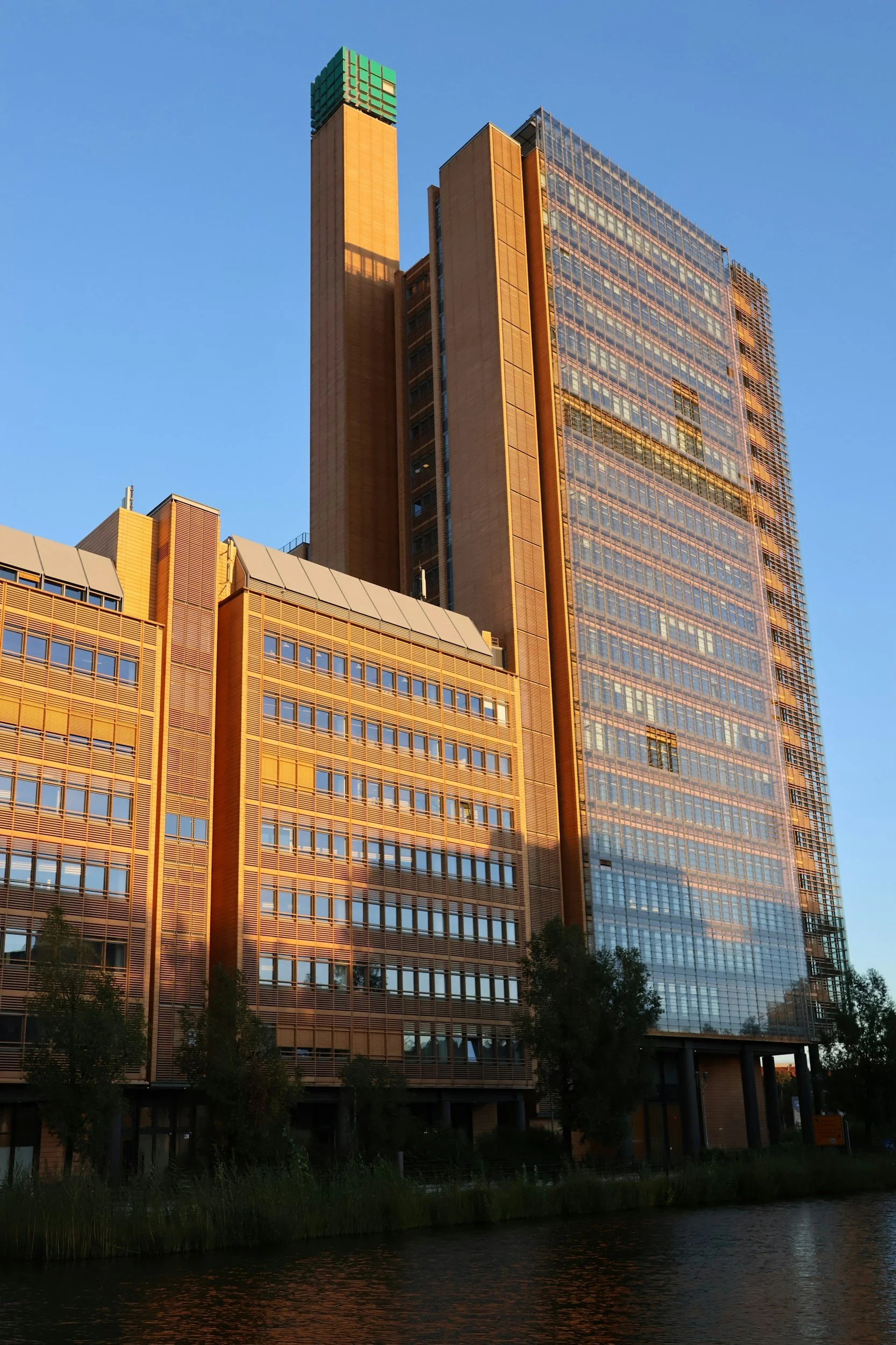 A modern high-rise building illuminated by sunlight, with reflective glass windows, next to a water body and trees at the bottom