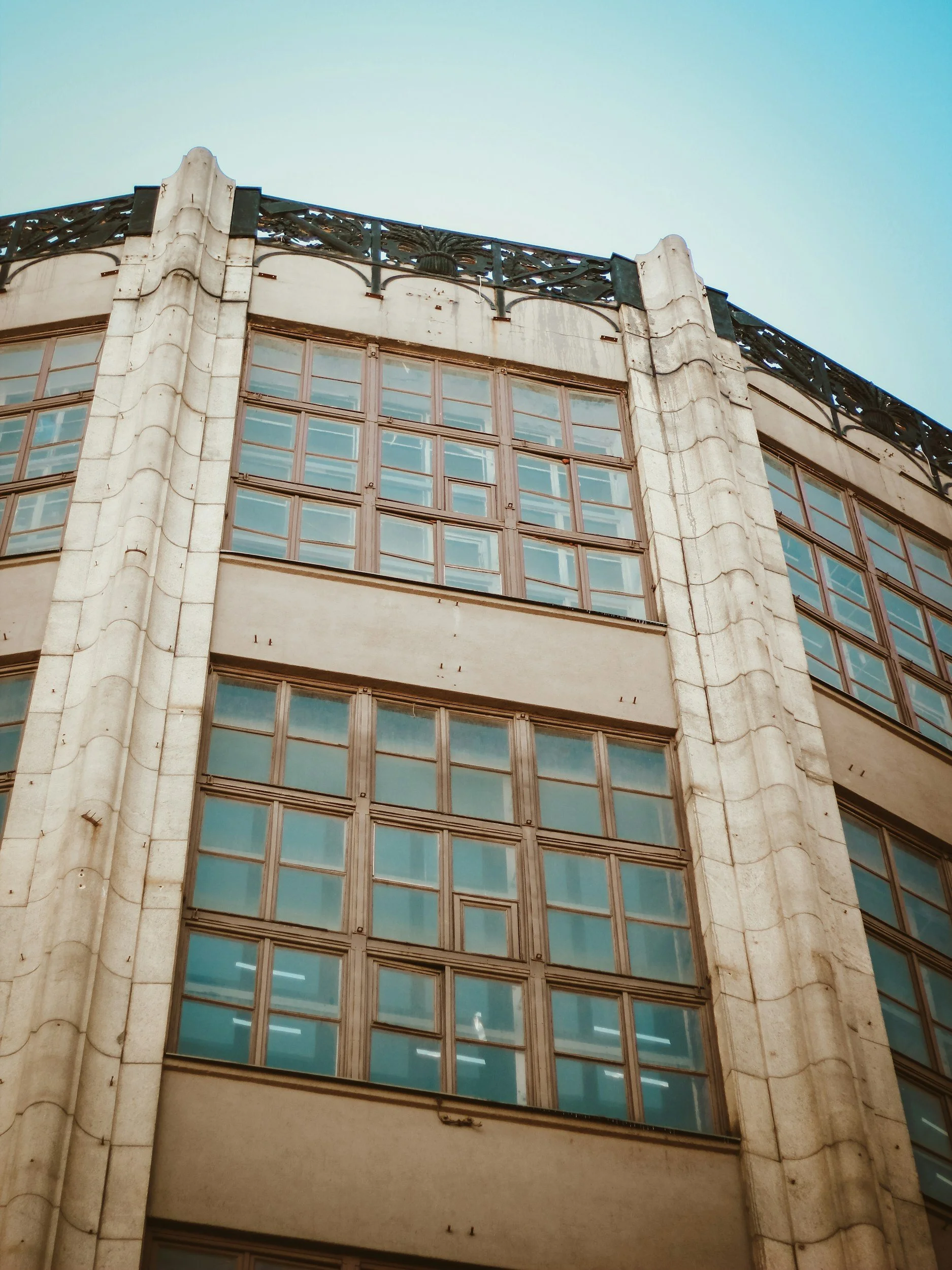 A close-up view of a tall, vintage, multi-story building with large windows and a decorative iron railing on the roof, set against a clear blue sky.