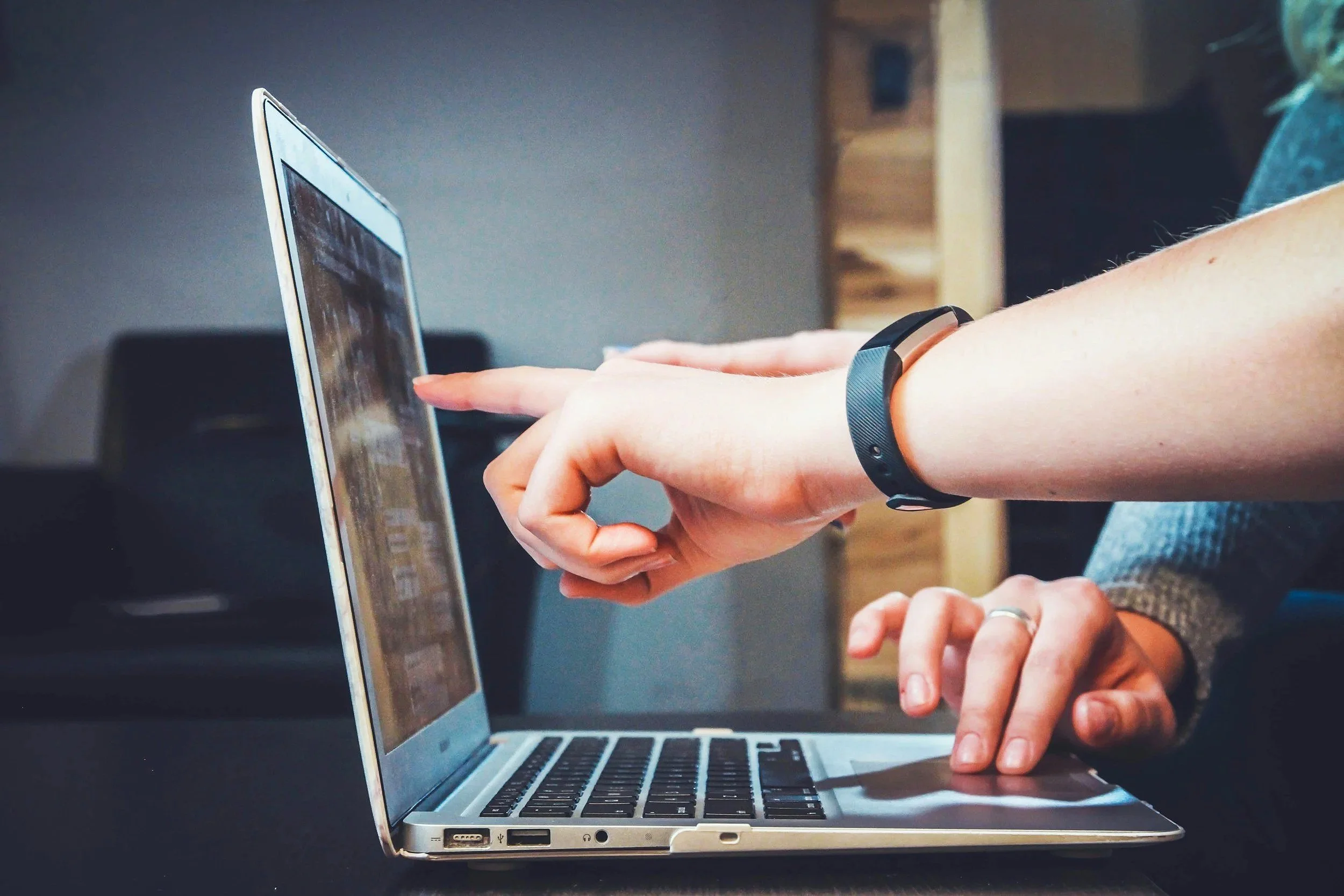 Person using a laptop computer with one hand, wearing a fitness tracker on their wrist, sitting at a dark table.
