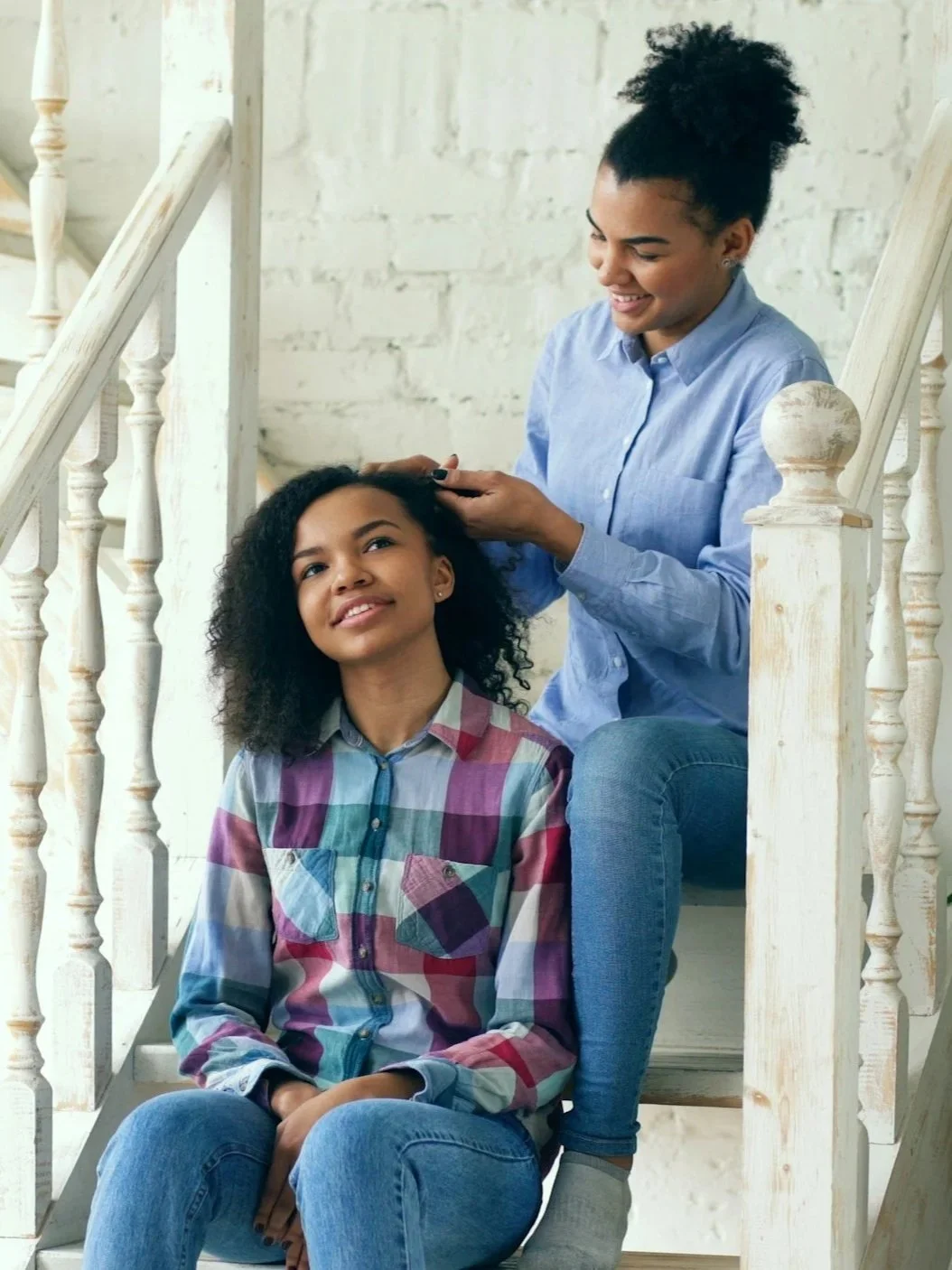 A woman combing a young girl's hair while they sit on stairs in a bright room with white brick walls.