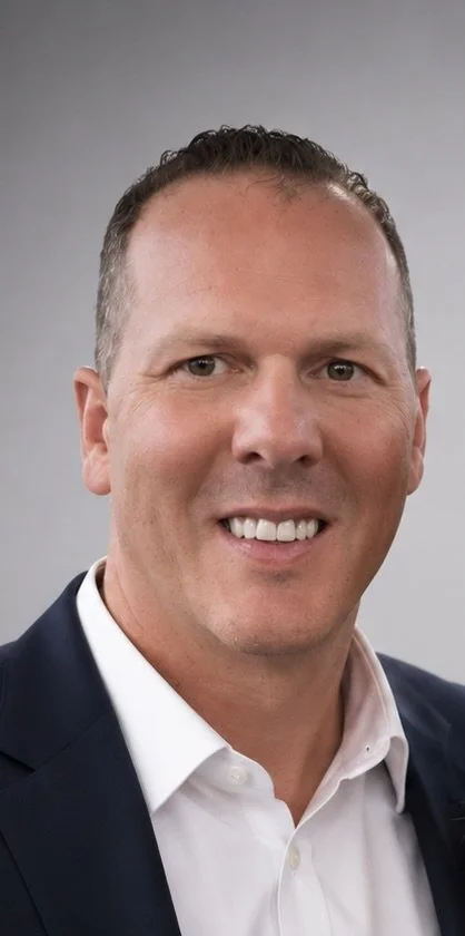 A smiling man with short dark hair, wearing a white dress shirt and a dark blazer, against a neutral background.