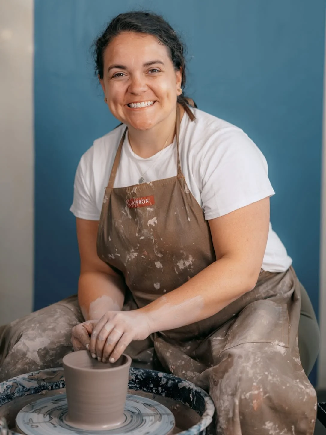 Meghann at her pottery wheel in her studio smiling at the camera.