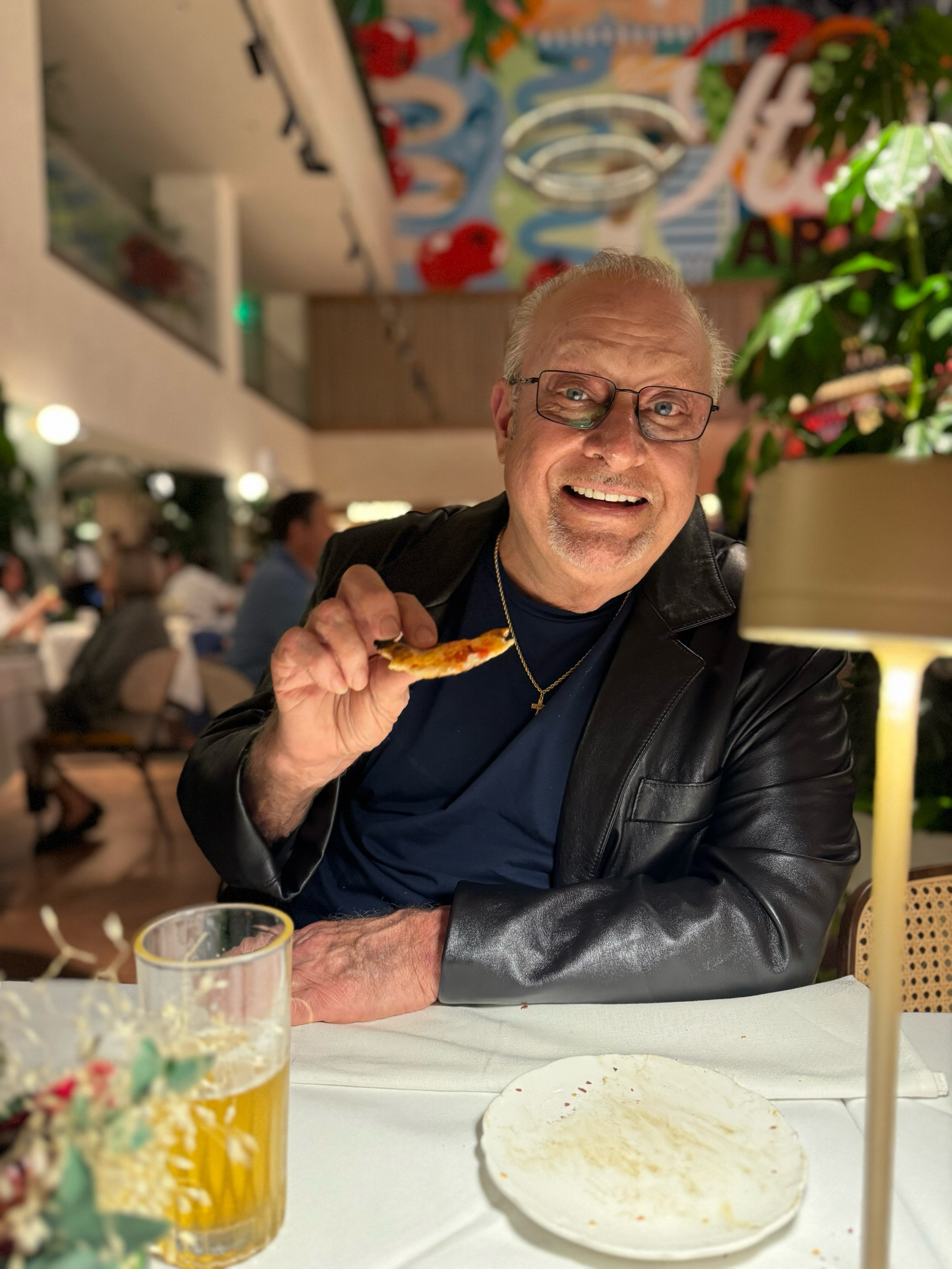 An elderly man with glasses smiling and holding a slice of pizza in a restaurant. The scene includes a table with an empty plate, a glass of beer, and a decorative Easter-themed cup, with other patrons and colorful artwork on the wall in the background.