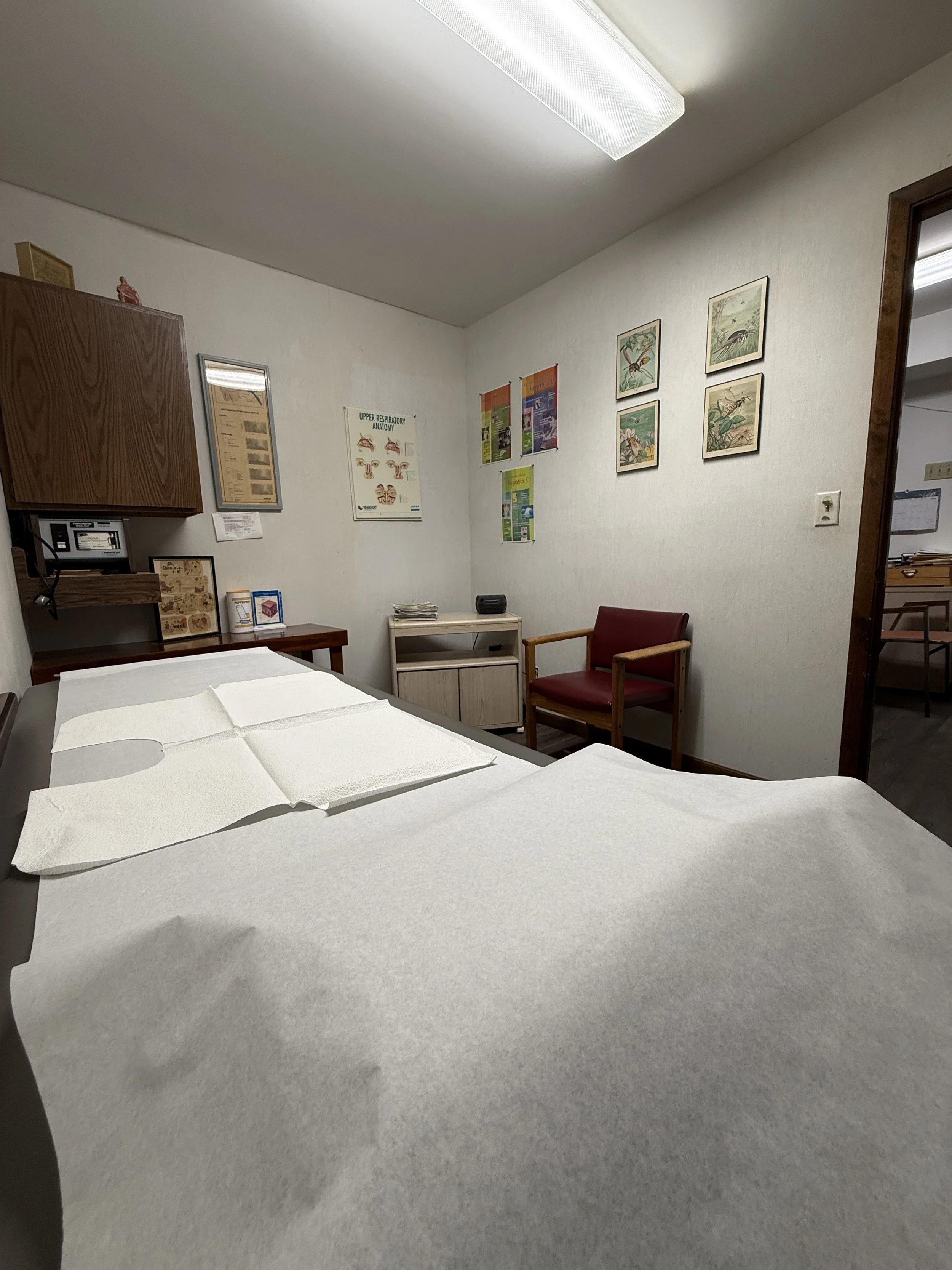 A medical examination room with a table covered in white paper, a wooden cabinet, framed medical posters, a small white side table, a red armchair, and a doorway leading to another room.