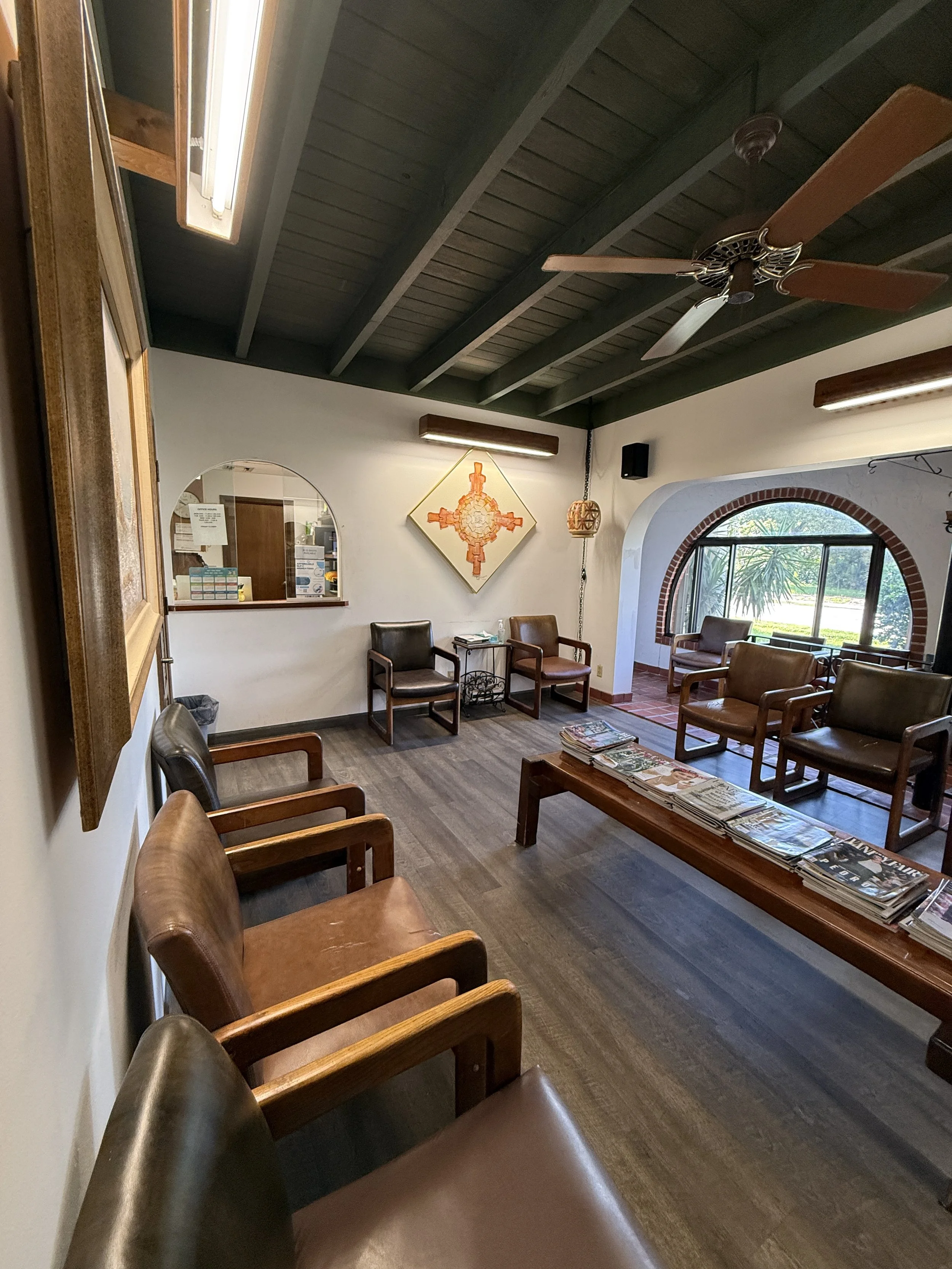 A waiting room with several brown leather chairs arranged along the walls, a wooden coffee table with magazines, a small side table, a decorative wall hanging, a window with an arch, and a view of green plants outside. The ceiling has dark painted wooden beams and a ceiling fan.