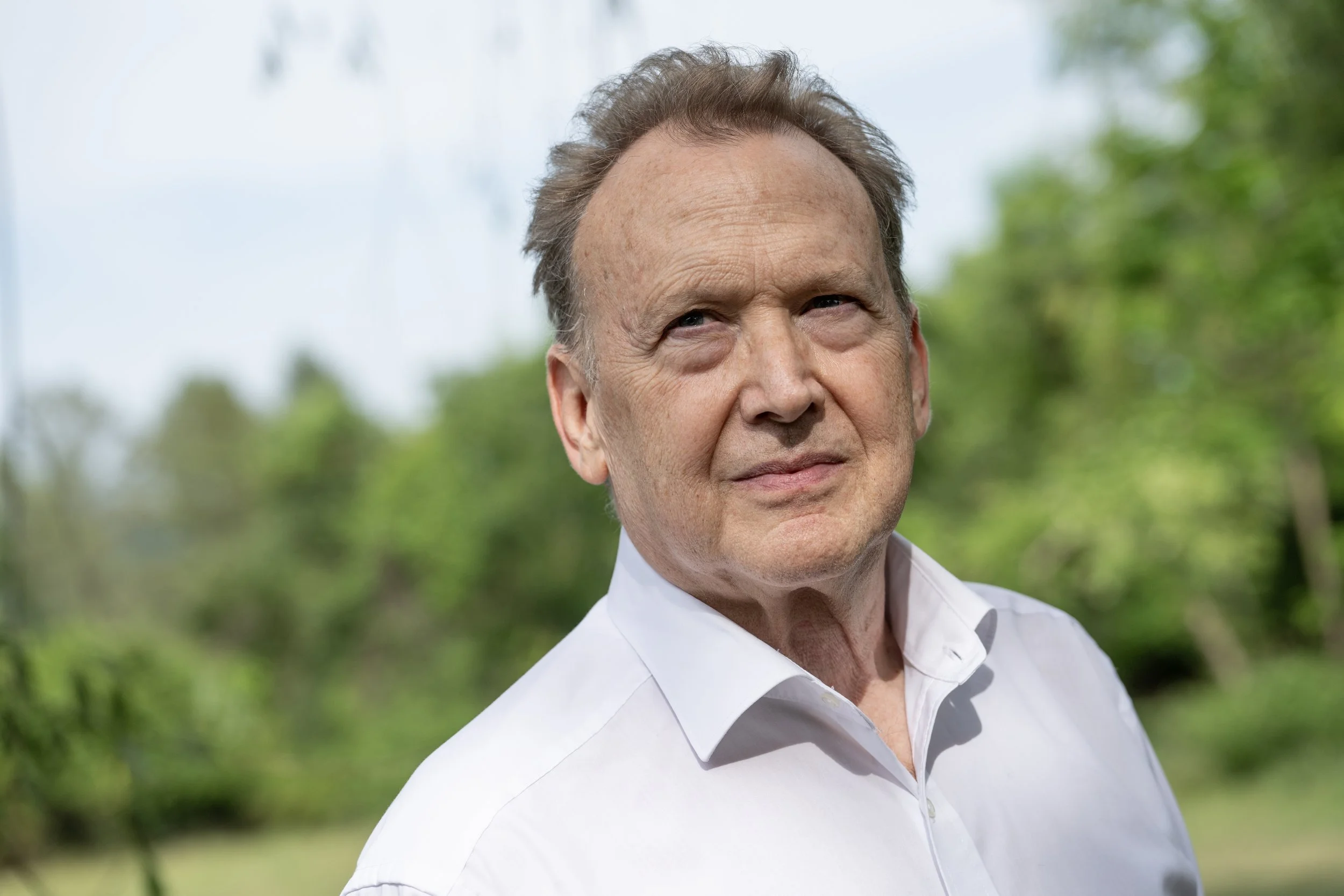 Close-up of Peter Orth, wearing a white button-up shirt, standing outdoors with blurred green foliage in the background.