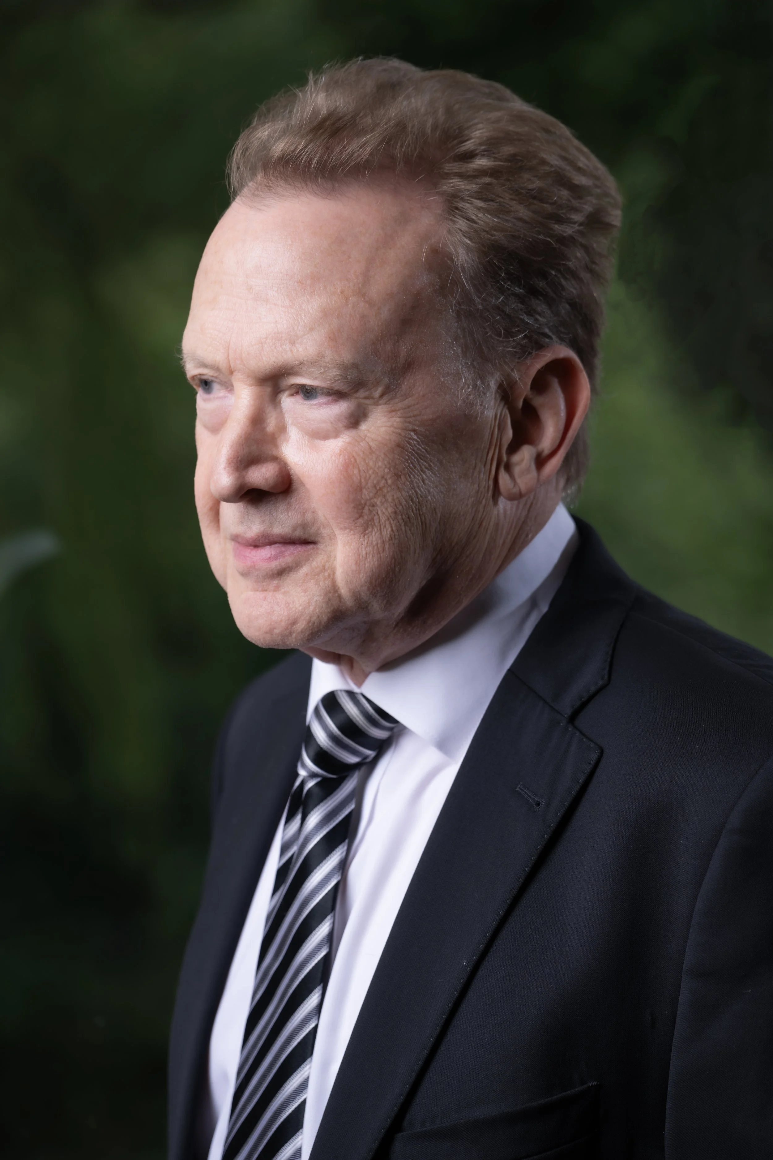 Close-up of Peter Orth, wearing a black tuxedo and a striped black and white tie, outdoors with blurred green foliage in the background.
