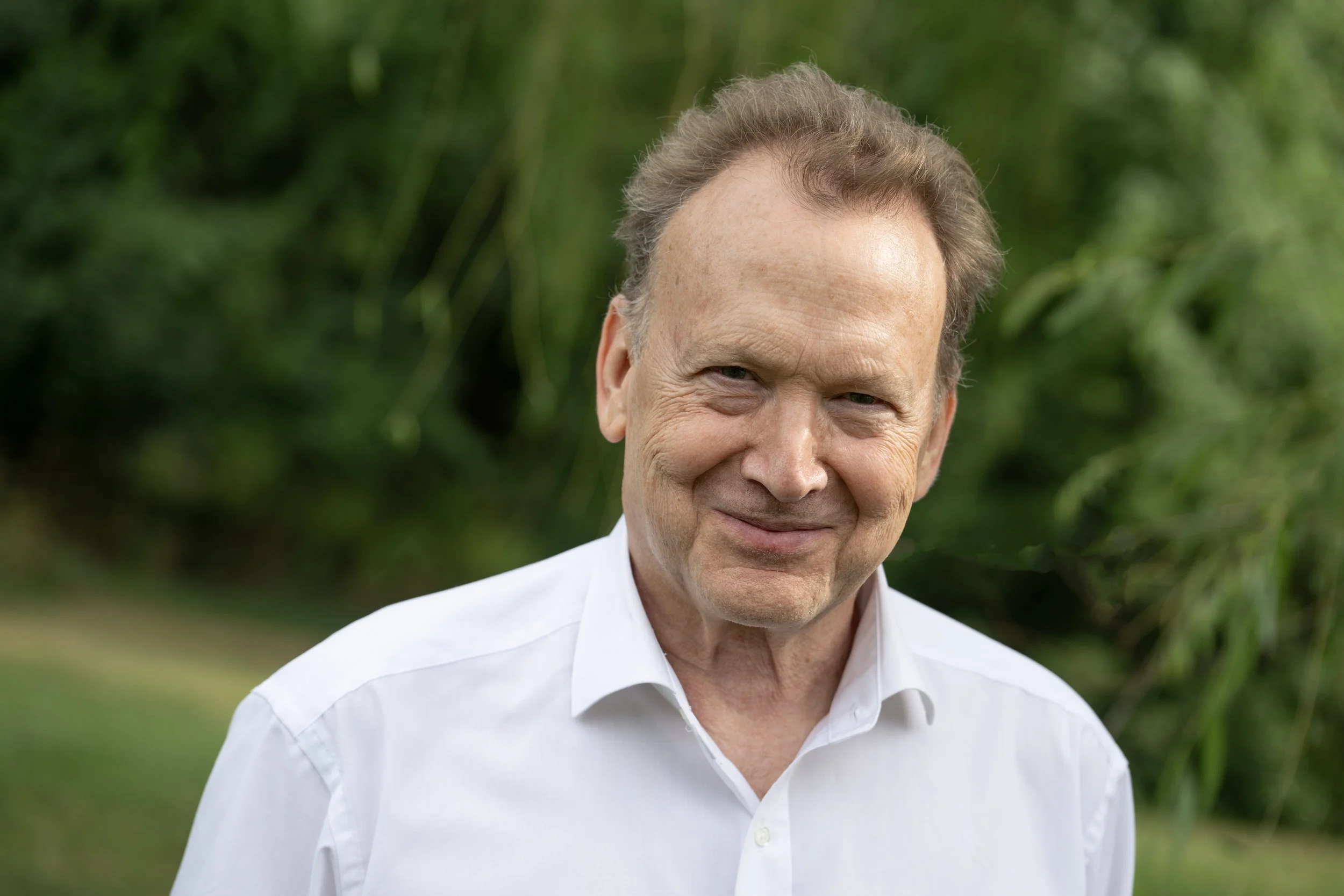Peter Orth, smiling, wearing a white collared shirt, standing outdoors with a lush green background.