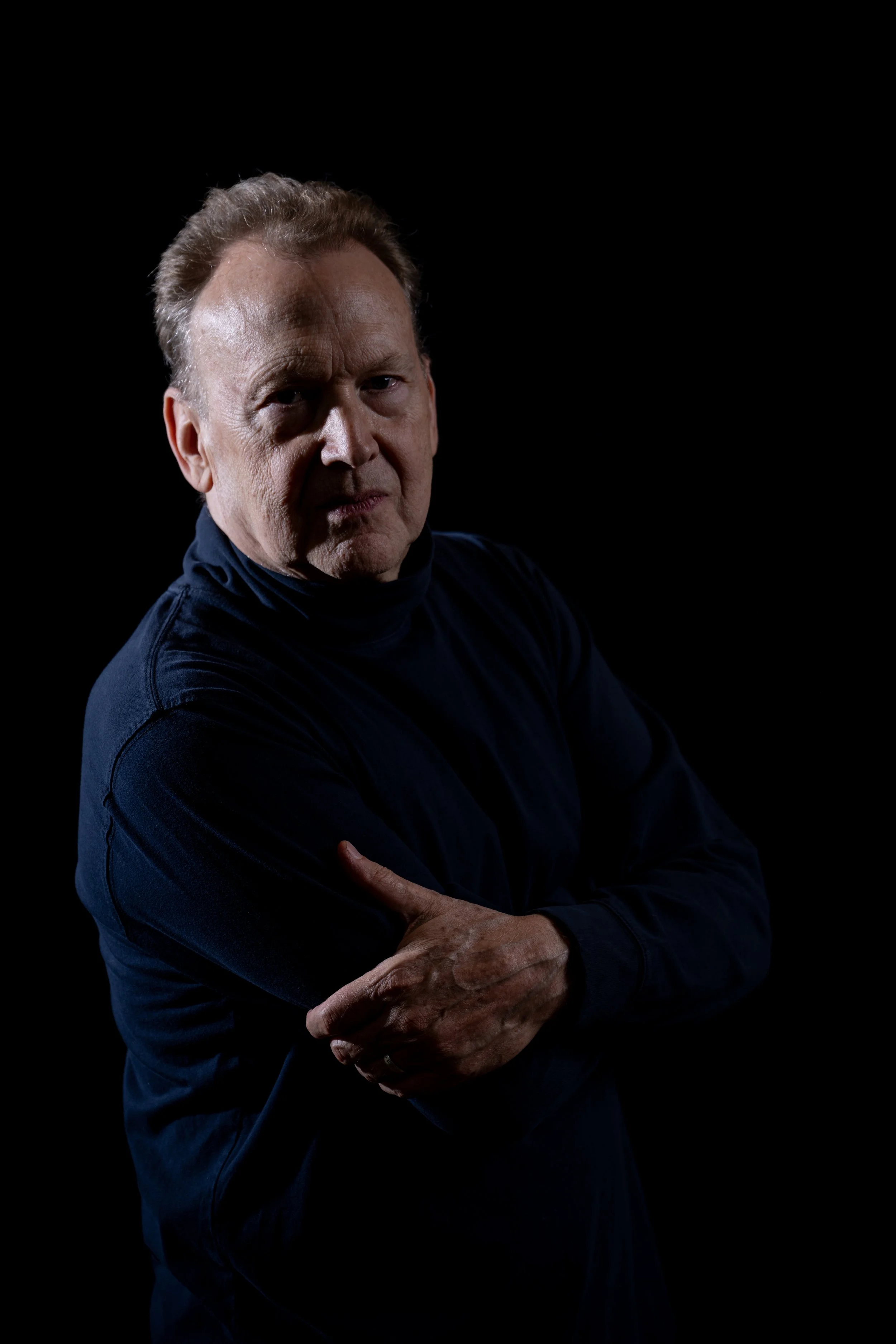 Peter Orth wearing a dark long-sleeve shirt, crossing his arms, posed against a black background.