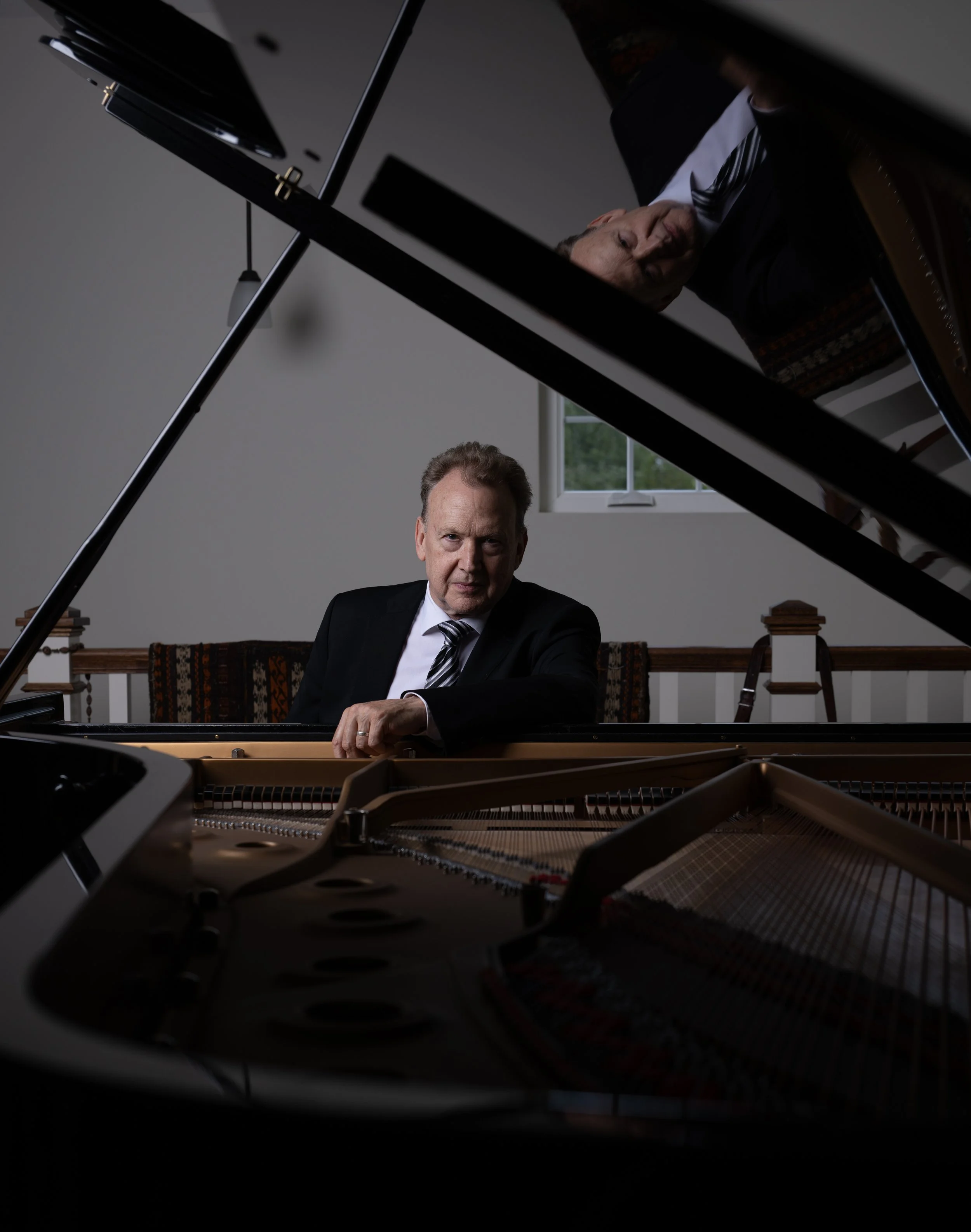 Peter Orth in a suit and tie sitting at a grand piano, with his face reflected on the open piano lid. The room has a window in the background and a wooden bench with books.