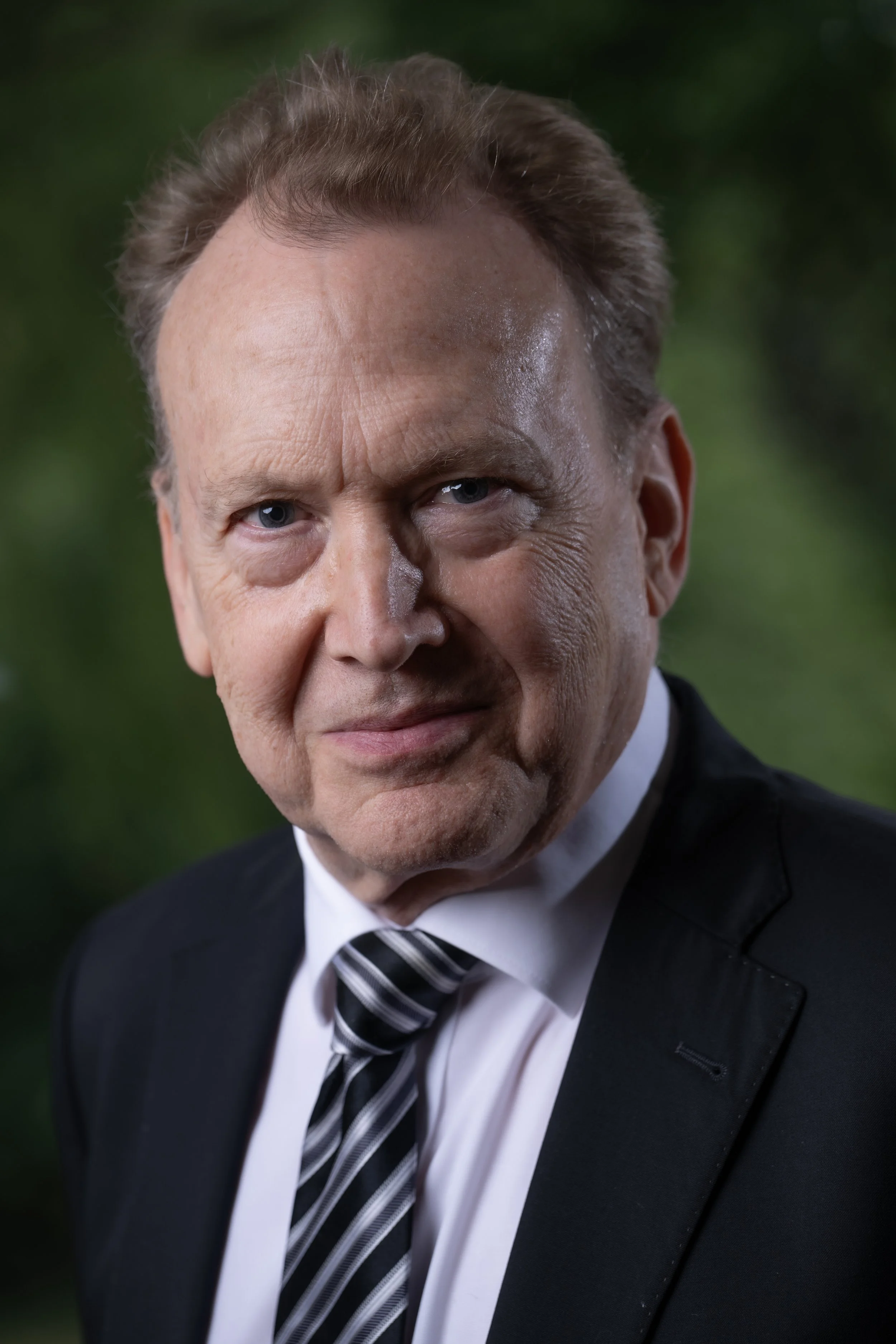 Close-up of Peter Orth in formal attire, wearing a tuxedo with a striped tie, standing outdoors with a blurred green background.