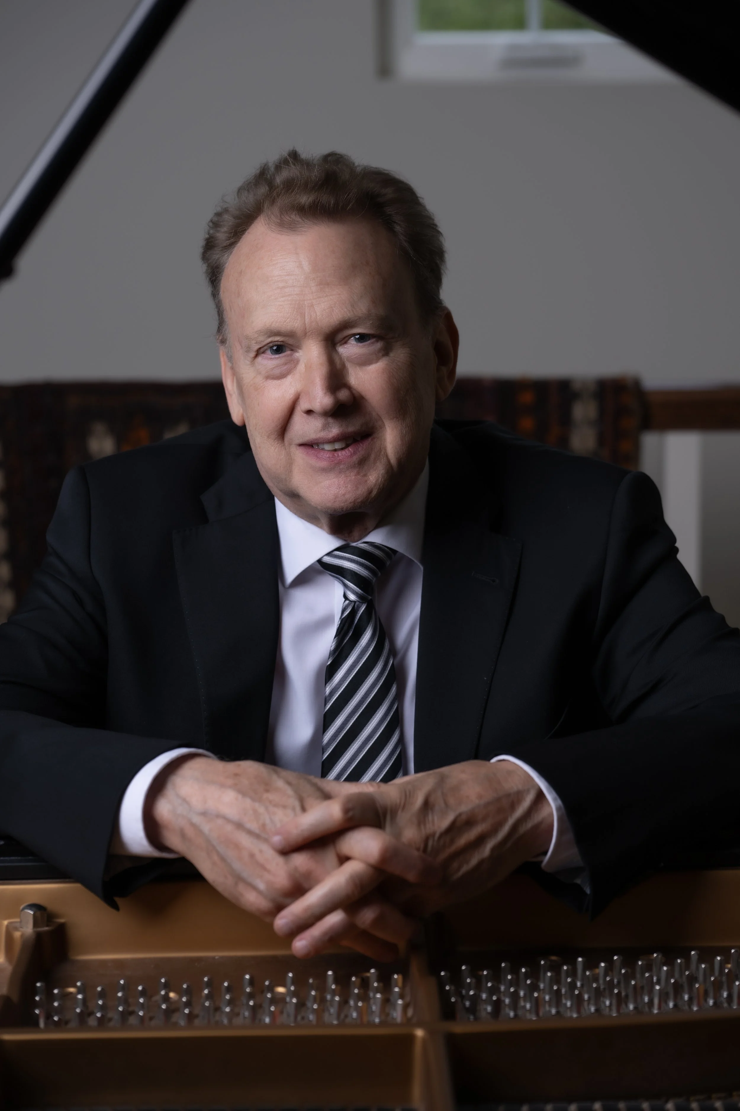 Peter Orth in a black suit and striped tie sitting at a piano, with his hands clasped on the piano's open lid, smiling at the camera.