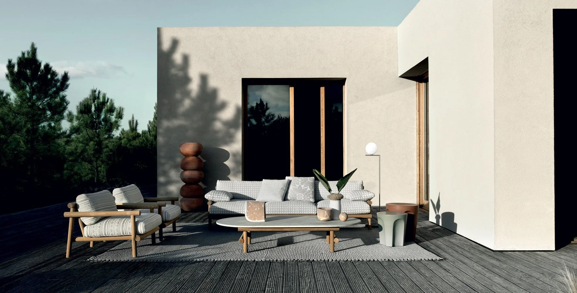 Modern outdoor patio with seating area including a sofa and chairs, a round coffee table, various decorative vases, a tall brown sculpture, a white spherical lamp, and plants, with a backdrop of green trees and a minimalist building wall.